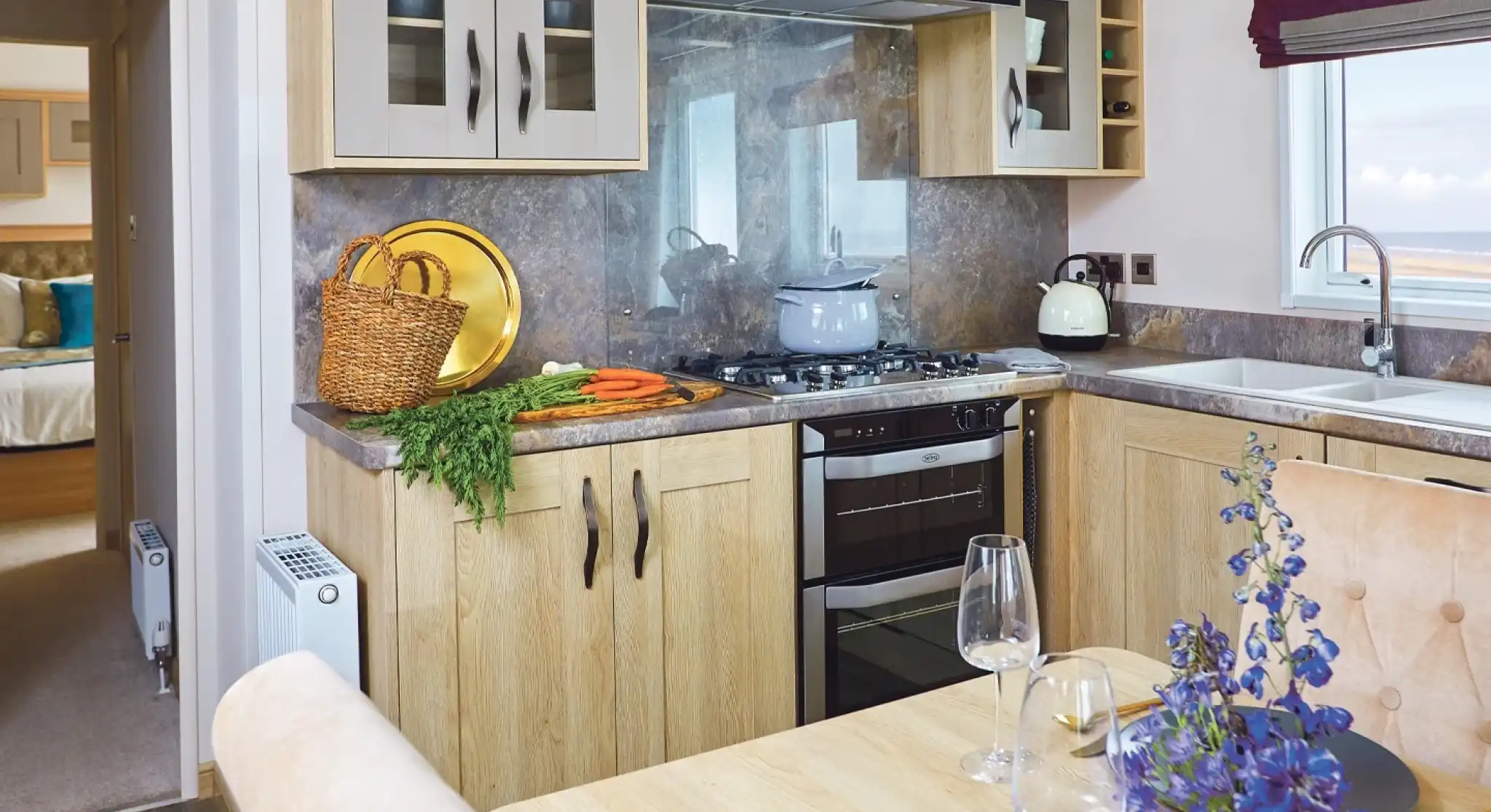 A modern kitchen with light wood cabinetry, a gas stove, and a countertop. A dining table is set with plates and wine glasses, while a bouquet of purple flowers adds a decorative touch. Fresh vegetables are placed on the counter, and a view of the outdoors is visible through a window.