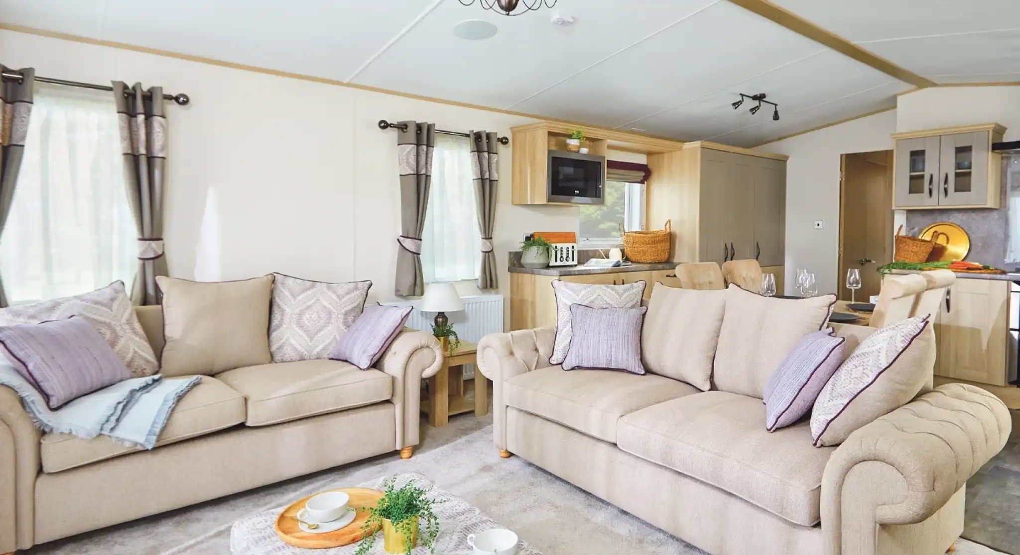 A cozy living room featuring two beige sofas adorned with decorative pillows, a light-colored coffee table with snacks, and large windows draped with elegant curtains. The room has a modern decor style with soft lighting and an open layout leading to a kitchen area.