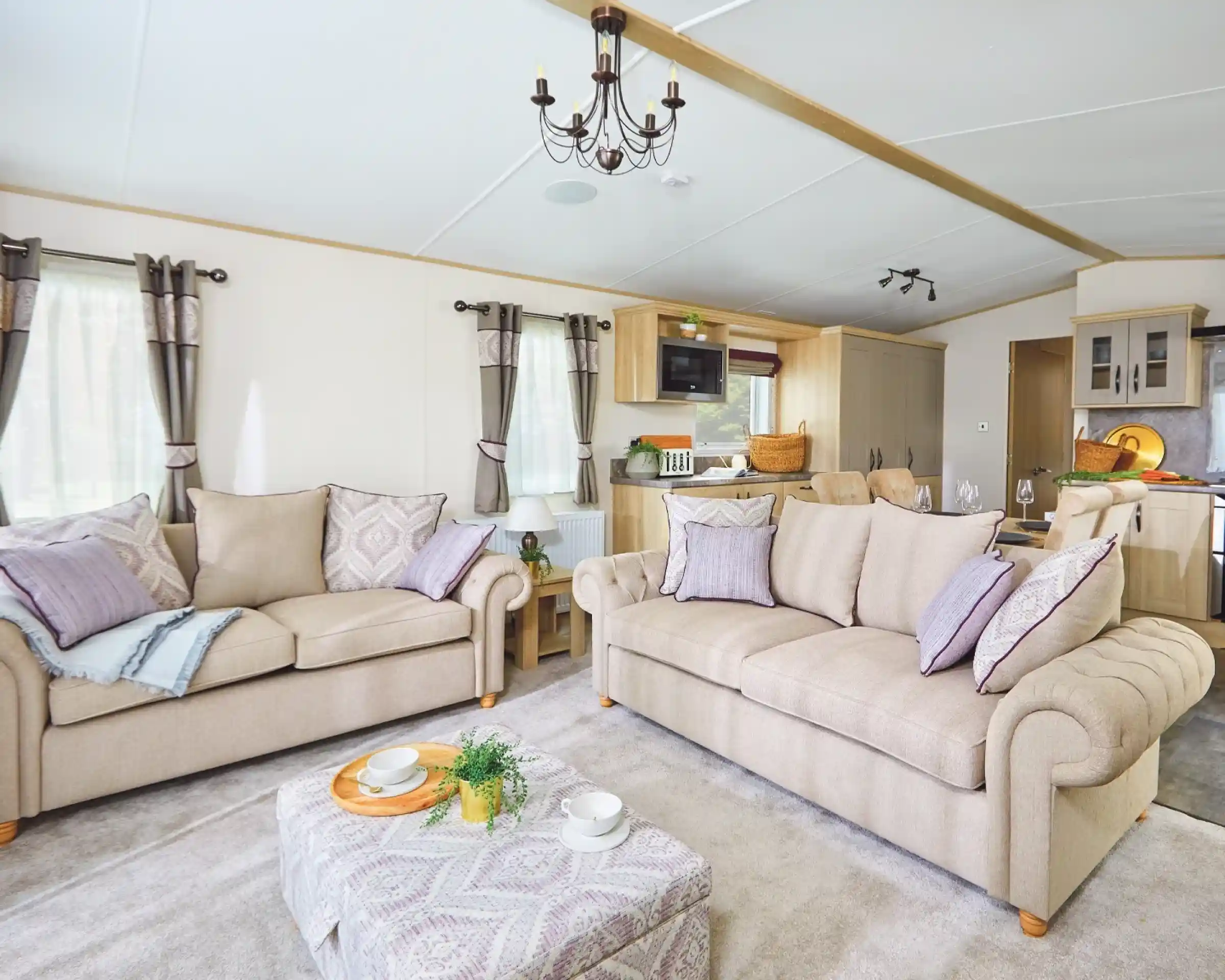 A cozy living room featuring two beige sofas adorned with decorative pillows, a light-colored coffee table with snacks, and large windows draped with elegant curtains. The room has a modern decor style with soft lighting and an open layout leading to a kitchen area.