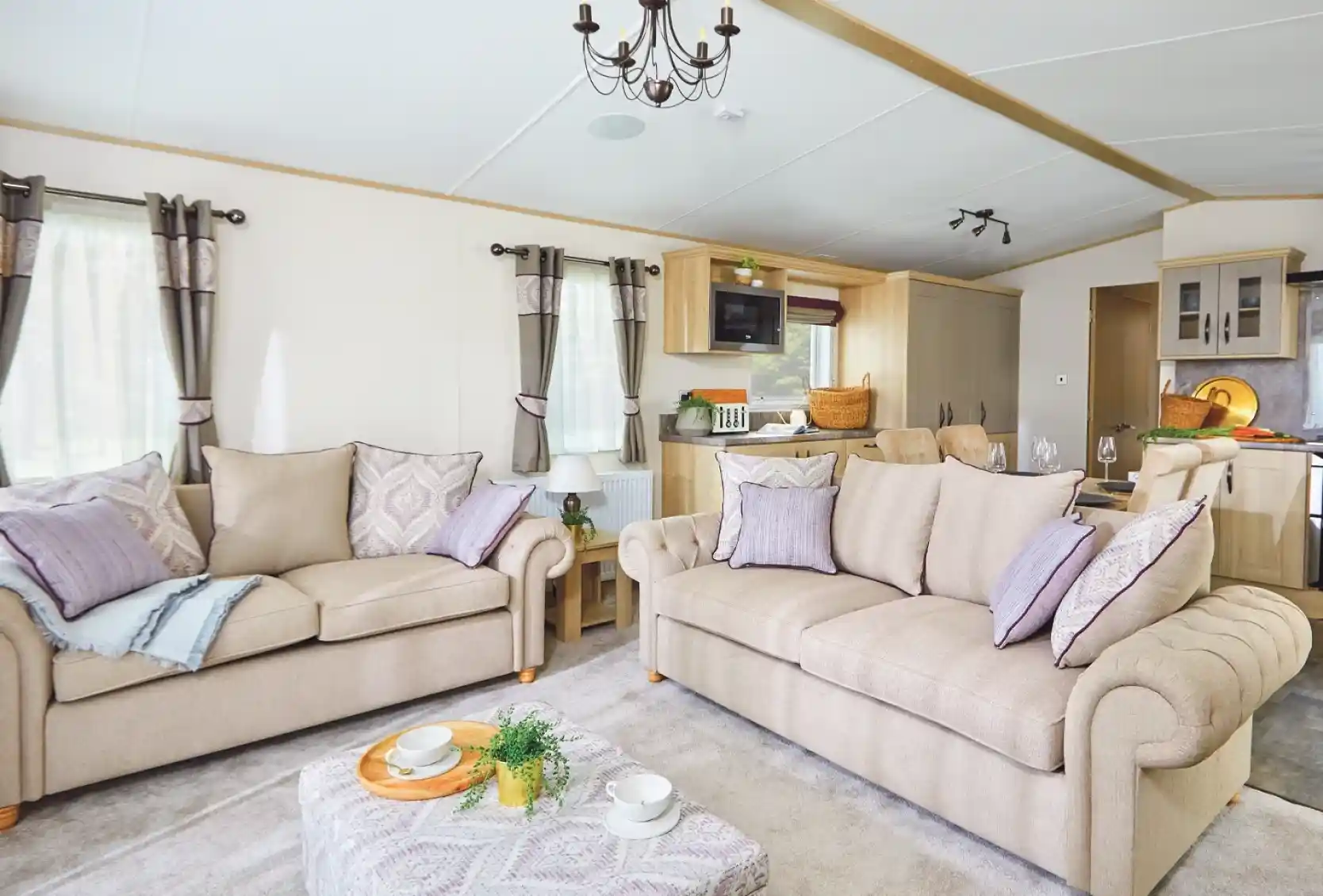 A cozy living room featuring two beige sofas adorned with decorative pillows, a light-colored coffee table with snacks, and large windows draped with elegant curtains. The room has a modern decor style with soft lighting and an open layout leading to a kitchen area.