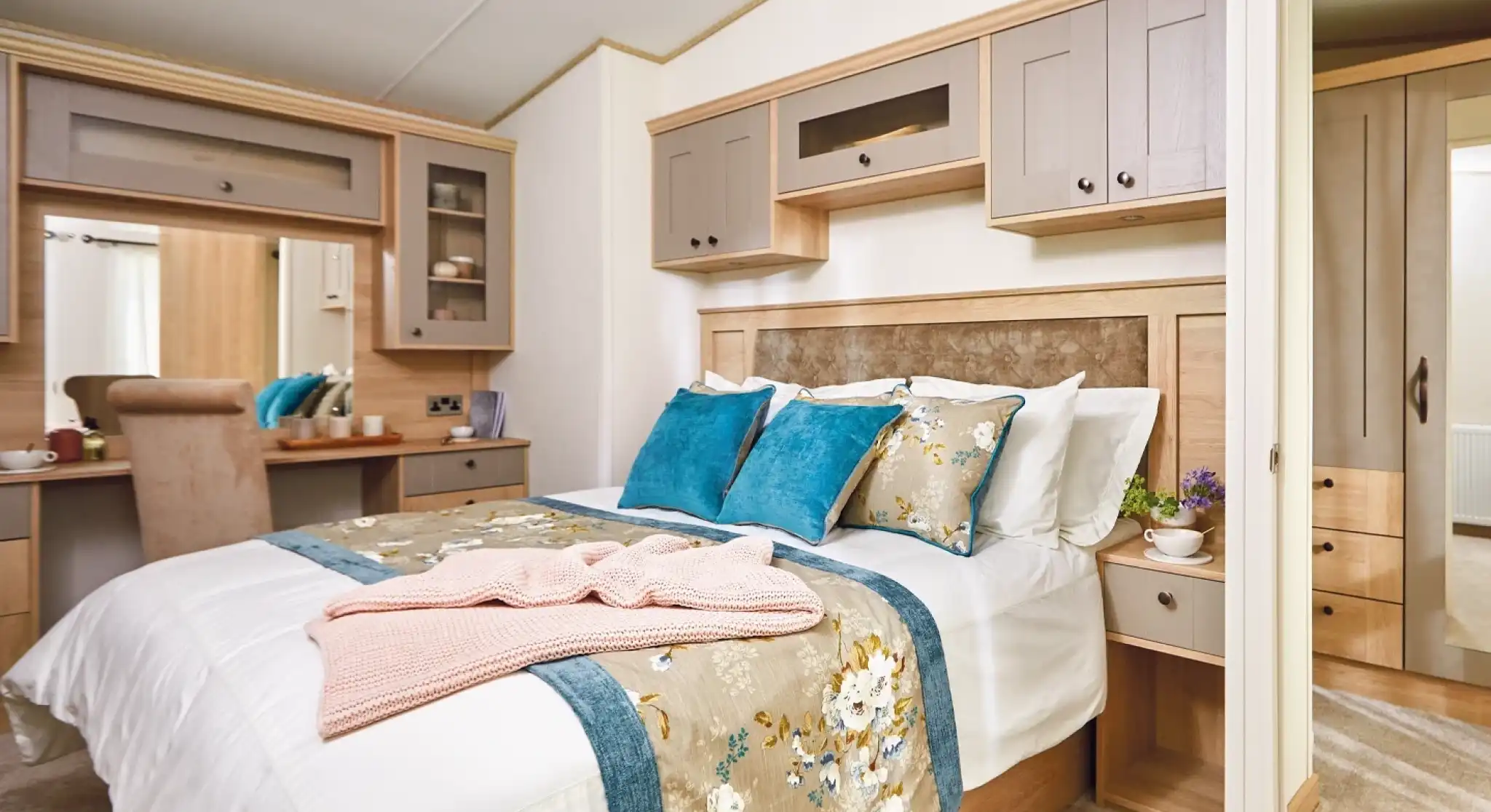 A cozy bedroom featuring a double bed with decorative pillows in shades of blue and white. The room has wooden cabinetry and a bedside table with a small plant. A plush blanket is draped over the bed, and there is a small desk area visible in the background. Light-colored walls enhance the warm atmosphere.