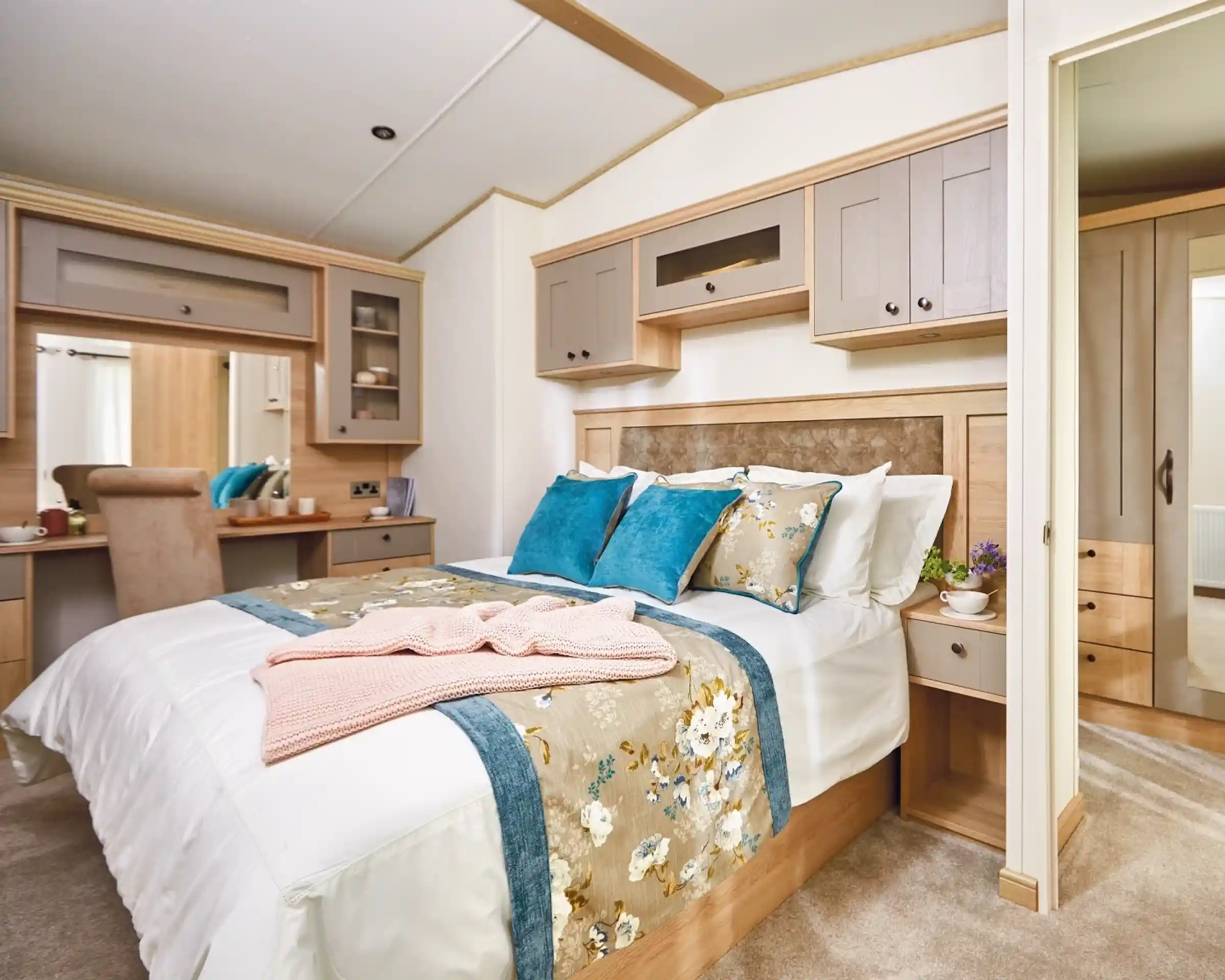 A cozy bedroom featuring a double bed with decorative pillows in shades of blue and white. The room has wooden cabinetry and a bedside table with a small plant. A plush blanket is draped over the bed, and there is a small desk area visible in the background. Light-colored walls enhance the warm atmosphere.