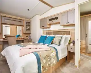 A cozy bedroom featuring a double bed with decorative pillows in shades of blue and white. The room has wooden cabinetry and a bedside table with a small plant. A plush blanket is draped over the bed, and there is a small desk area visible in the background. Light-colored walls enhance the warm atmosphere.