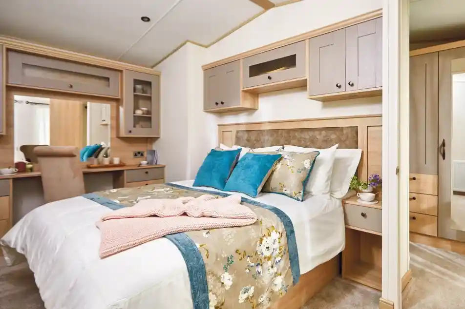 A cozy bedroom featuring a double bed with decorative pillows in shades of blue and white. The room has wooden cabinetry and a bedside table with a small plant. A plush blanket is draped over the bed, and there is a small desk area visible in the background. Light-colored walls enhance the warm atmosphere.