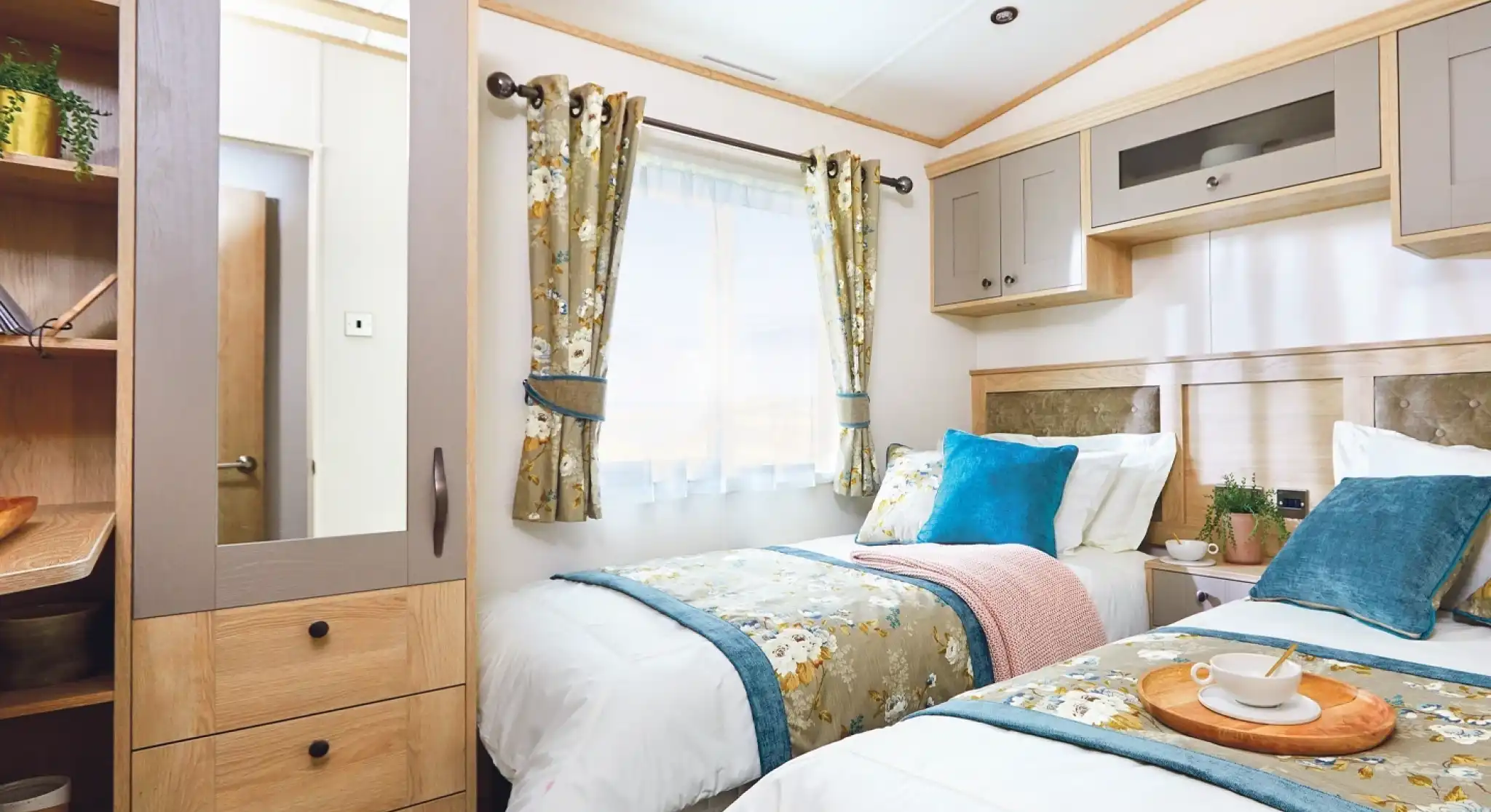 A cozy bedroom featuring two single beds with decorative pillows, a bedside table with a tray, and a window with floral curtains. A mirrored wardrobe is visible, along with soft lighting and neutral-colored walls.
