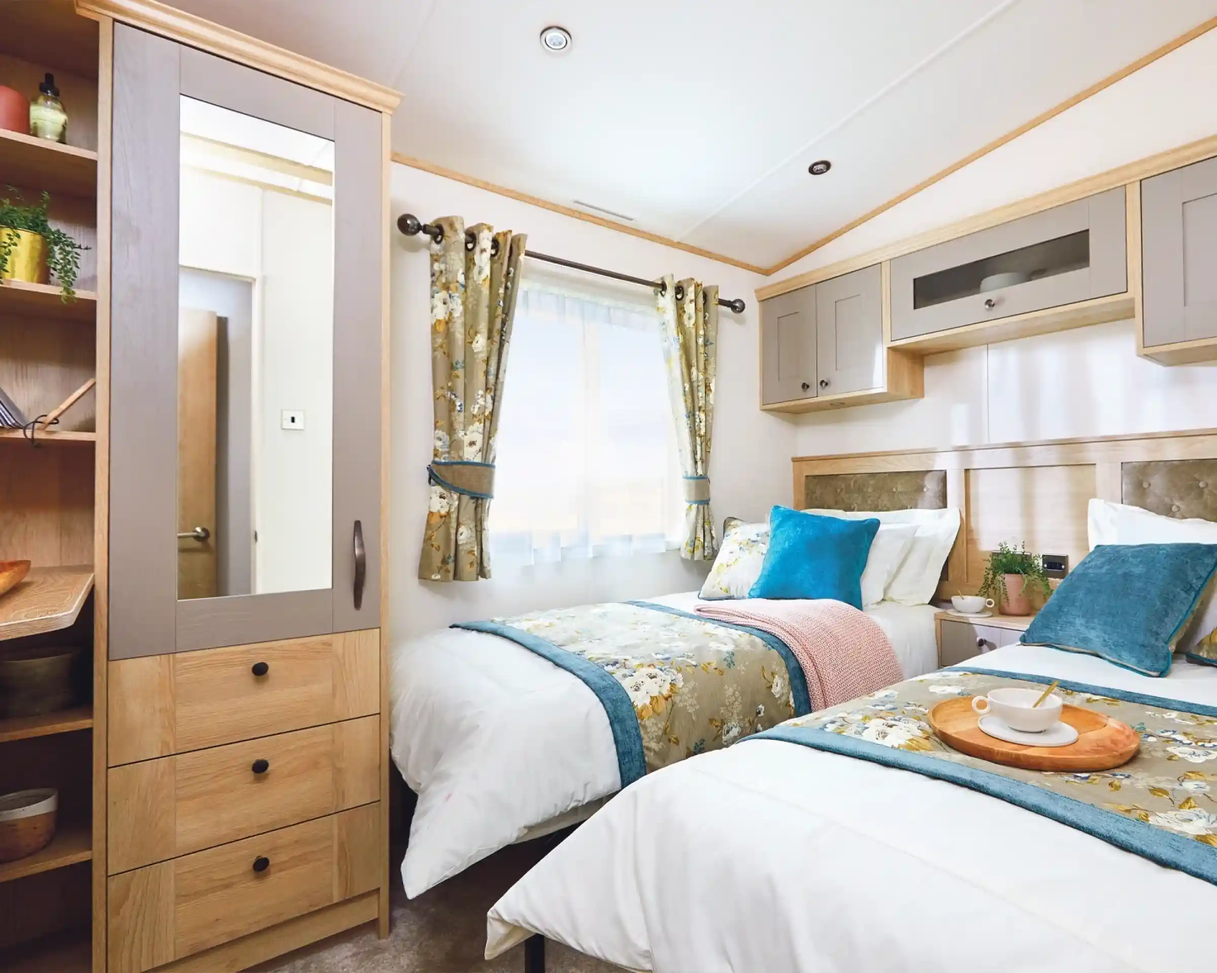 A cozy bedroom featuring two single beds with decorative pillows, a bedside table with a tray, and a window with floral curtains. A mirrored wardrobe is visible, along with soft lighting and neutral-colored walls.