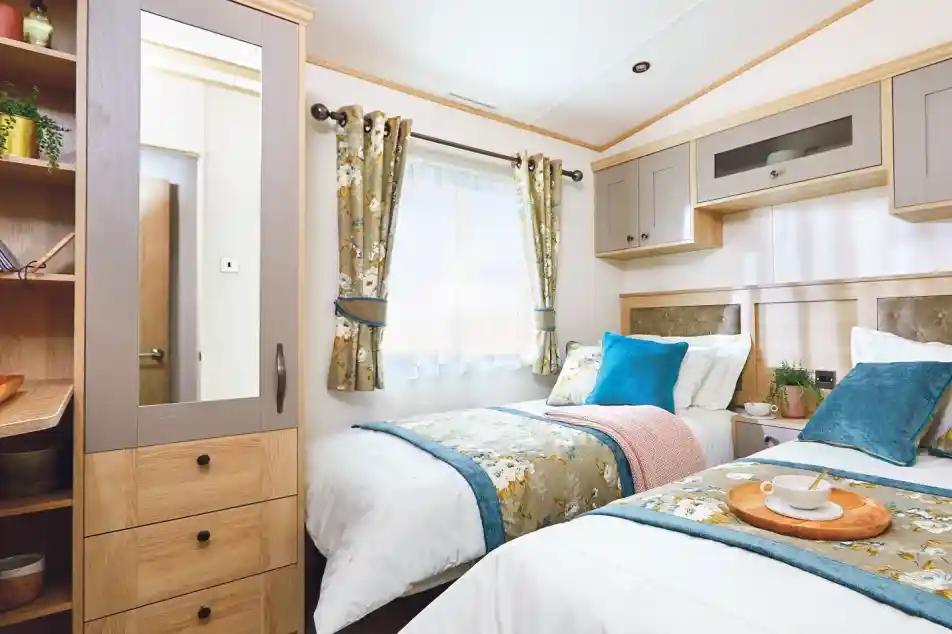 A cozy bedroom featuring two single beds with decorative pillows, a bedside table with a tray, and a window with floral curtains. A mirrored wardrobe is visible, along with soft lighting and neutral-colored walls.