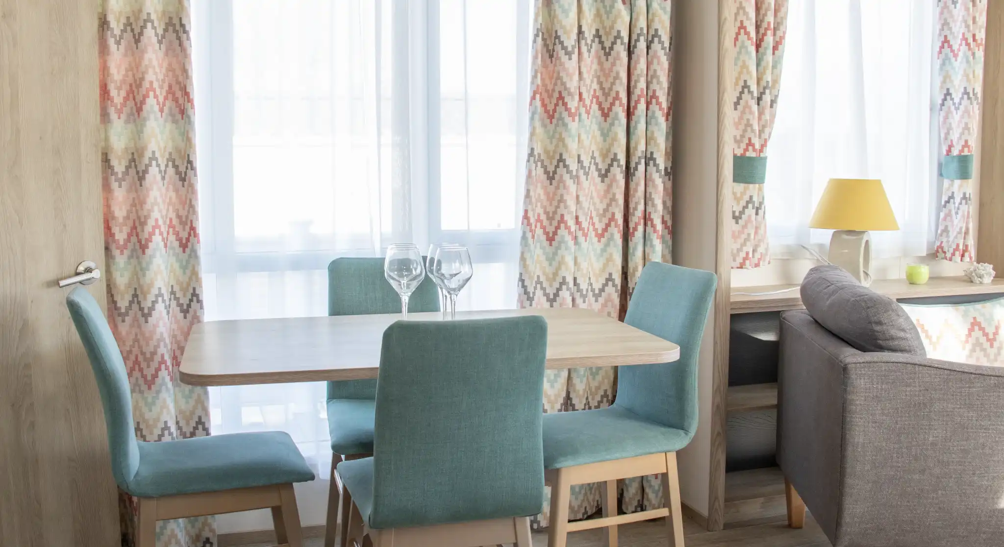 A cozy dining area featuring a wooden table surrounded by teal chairs. Bright curtains with a colorful zigzag pattern frame the windows, allowing natural light to fill the space. A lamp sits on a nearby side table next to a gray sofa, adding warmth to the room.