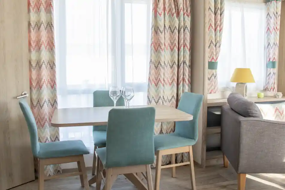 A cozy dining area featuring a wooden table surrounded by teal chairs. Bright curtains with a colorful zigzag pattern frame the windows, allowing natural light to fill the space. A lamp sits on a nearby side table next to a gray sofa, adding warmth to the room.