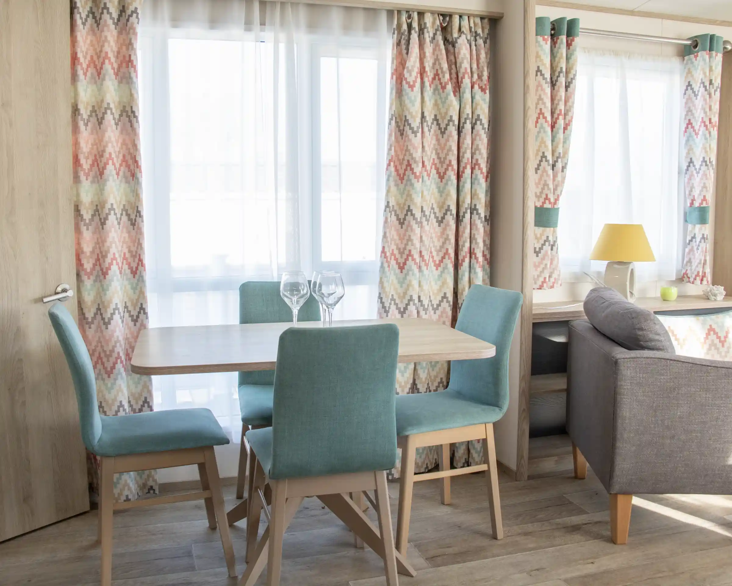 A cozy dining area featuring a wooden table surrounded by teal chairs. Bright curtains with a colorful zigzag pattern frame the windows, allowing natural light to fill the space. A lamp sits on a nearby side table next to a gray sofa, adding warmth to the room.
