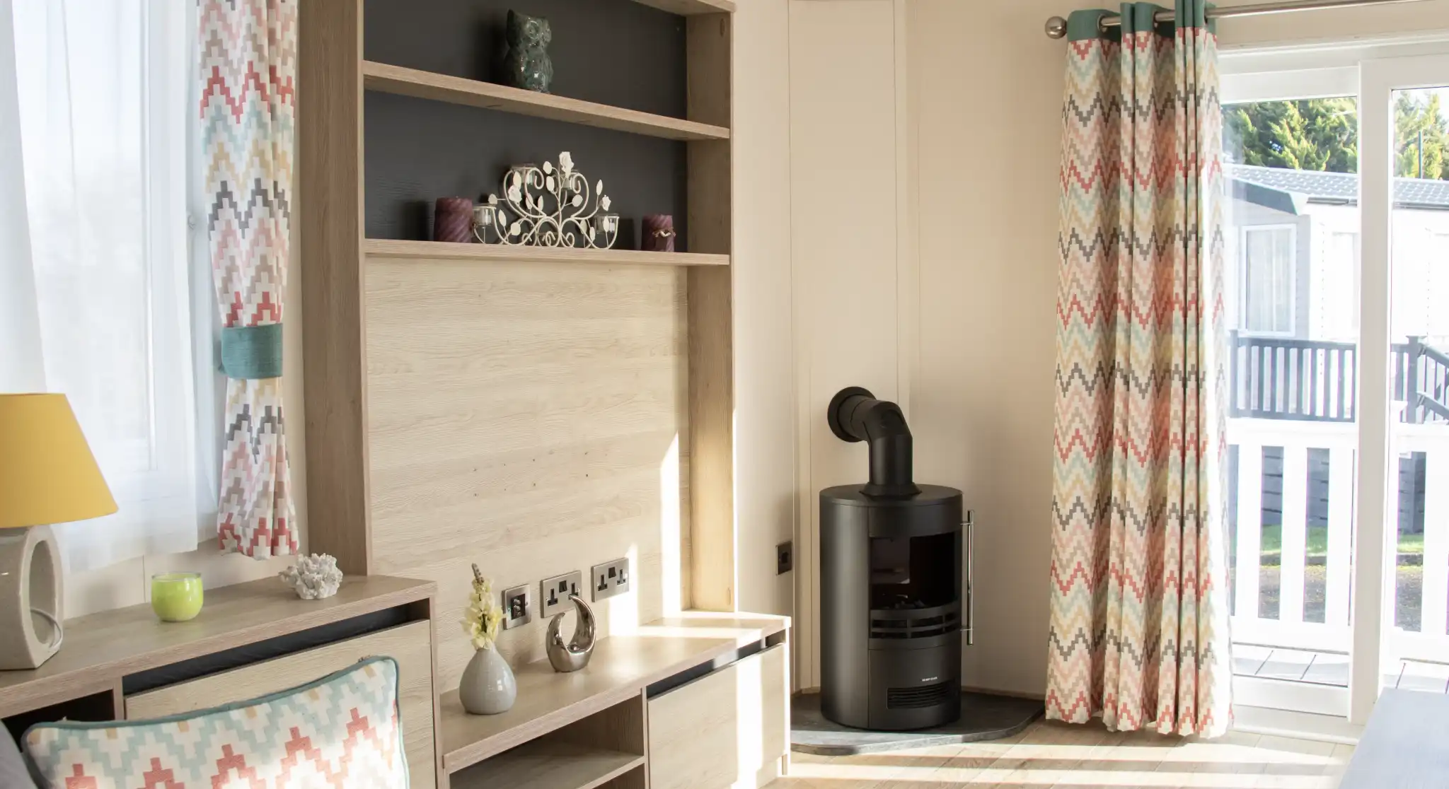 A cozy living room with light wood paneling, featuring a gray sofa and a decorative cushion. Colorful chevron-patterned curtains frame a window, and a modern black stove is positioned against one wall. A bookshelf with decorative items is mounted above a low wooden TV stand.