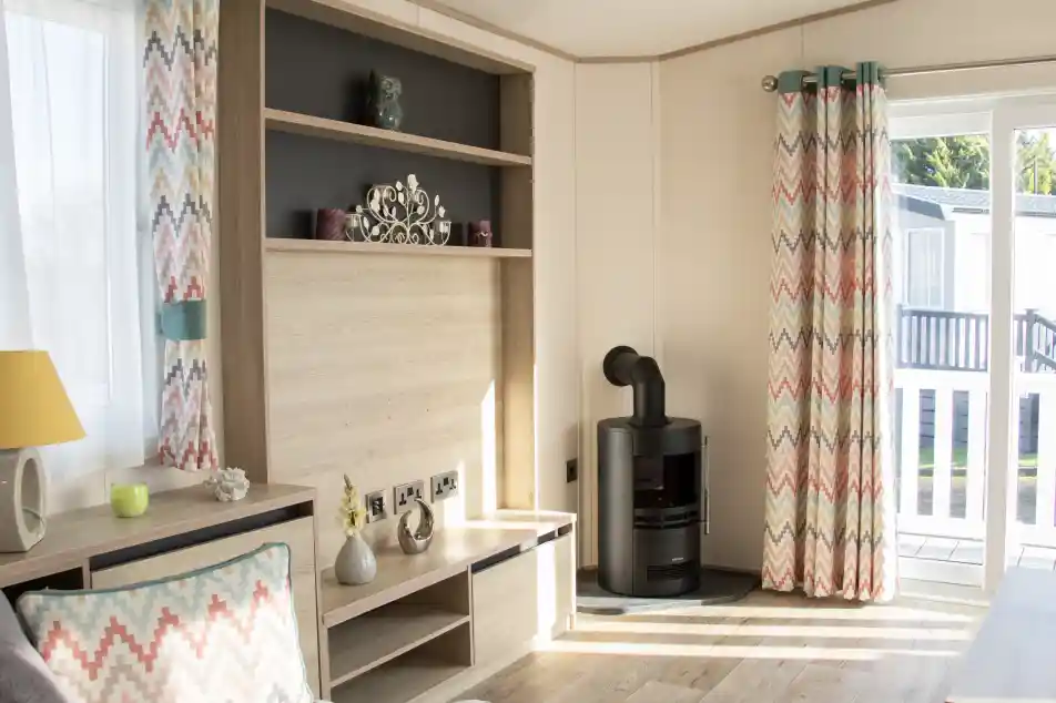 A cozy living room with light wood paneling, featuring a gray sofa and a decorative cushion. Colorful chevron-patterned curtains frame a window, and a modern black stove is positioned against one wall. A bookshelf with decorative items is mounted above a low wooden TV stand.