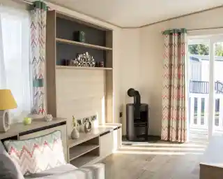 A cozy living room with light wood paneling, featuring a gray sofa and a decorative cushion. Colorful chevron-patterned curtains frame a window, and a modern black stove is positioned against one wall. A bookshelf with decorative items is mounted above a low wooden TV stand.