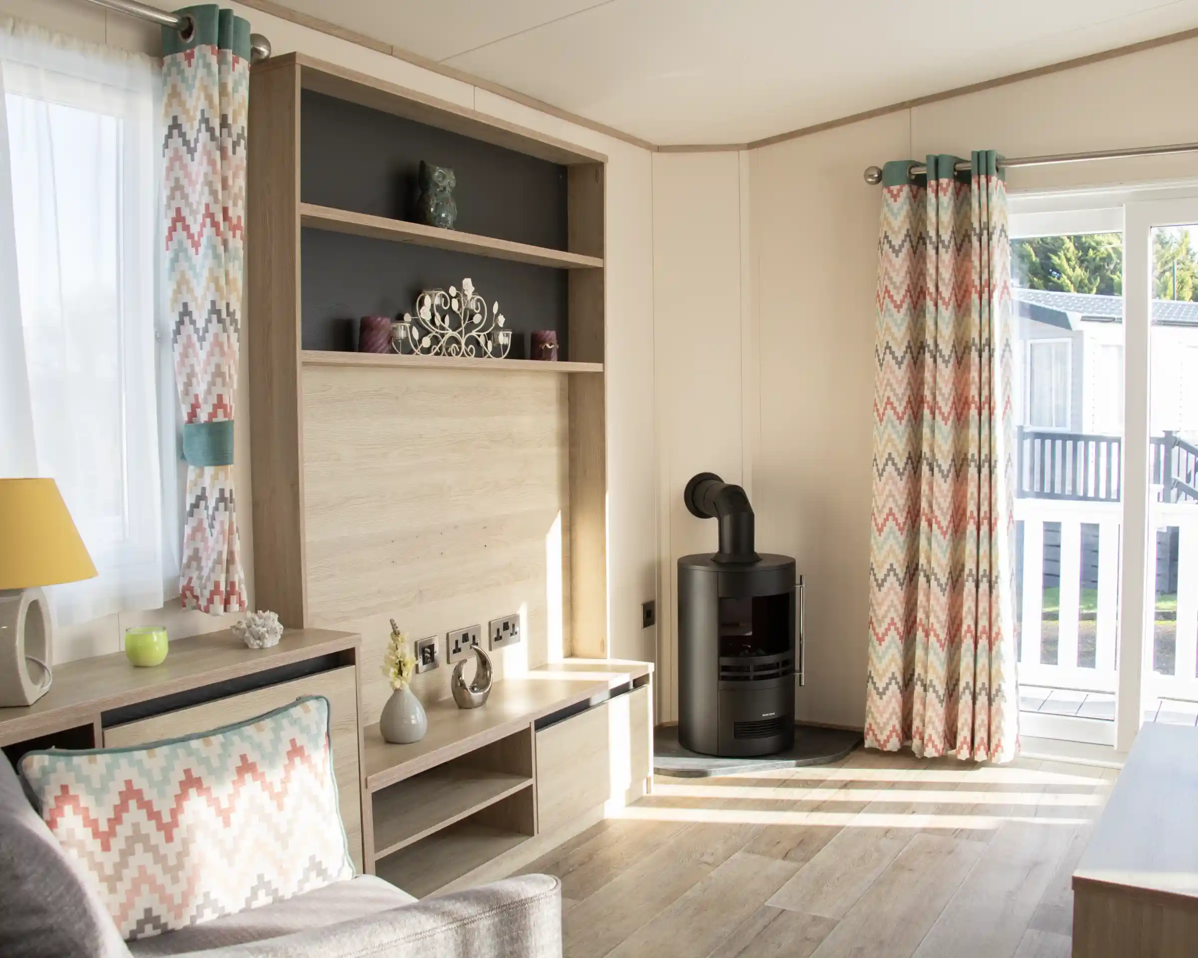 A cozy living room with light wood paneling, featuring a gray sofa and a decorative cushion. Colorful chevron-patterned curtains frame a window, and a modern black stove is positioned against one wall. A bookshelf with decorative items is mounted above a low wooden TV stand.