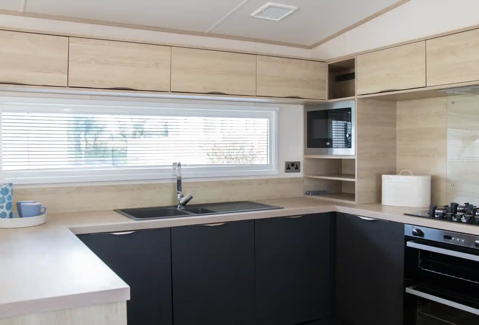 Modern kitchen featuring wooden cabinetry in light and dark tones. A large window with blinds provides natural light. The layout includes a sink, stovetop, and microwave, with minimalistic shelving above.