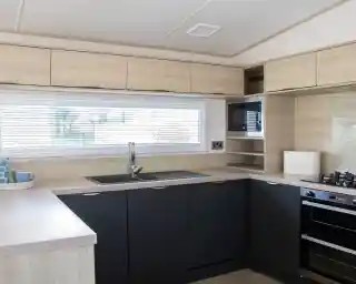 Modern kitchen featuring wooden cabinetry in light and dark tones. A large window with blinds provides natural light. The layout includes a sink, stovetop, and microwave, with minimalistic shelving above.
