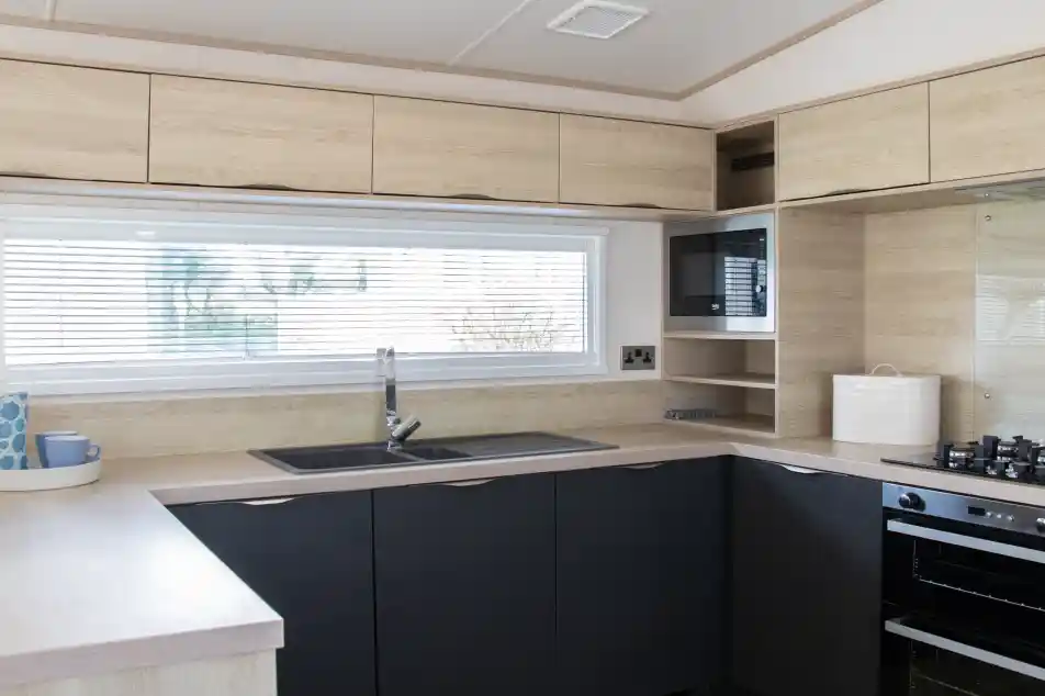 Modern kitchen featuring wooden cabinetry in light and dark tones. A large window with blinds provides natural light. The layout includes a sink, stovetop, and microwave, with minimalistic shelving above.