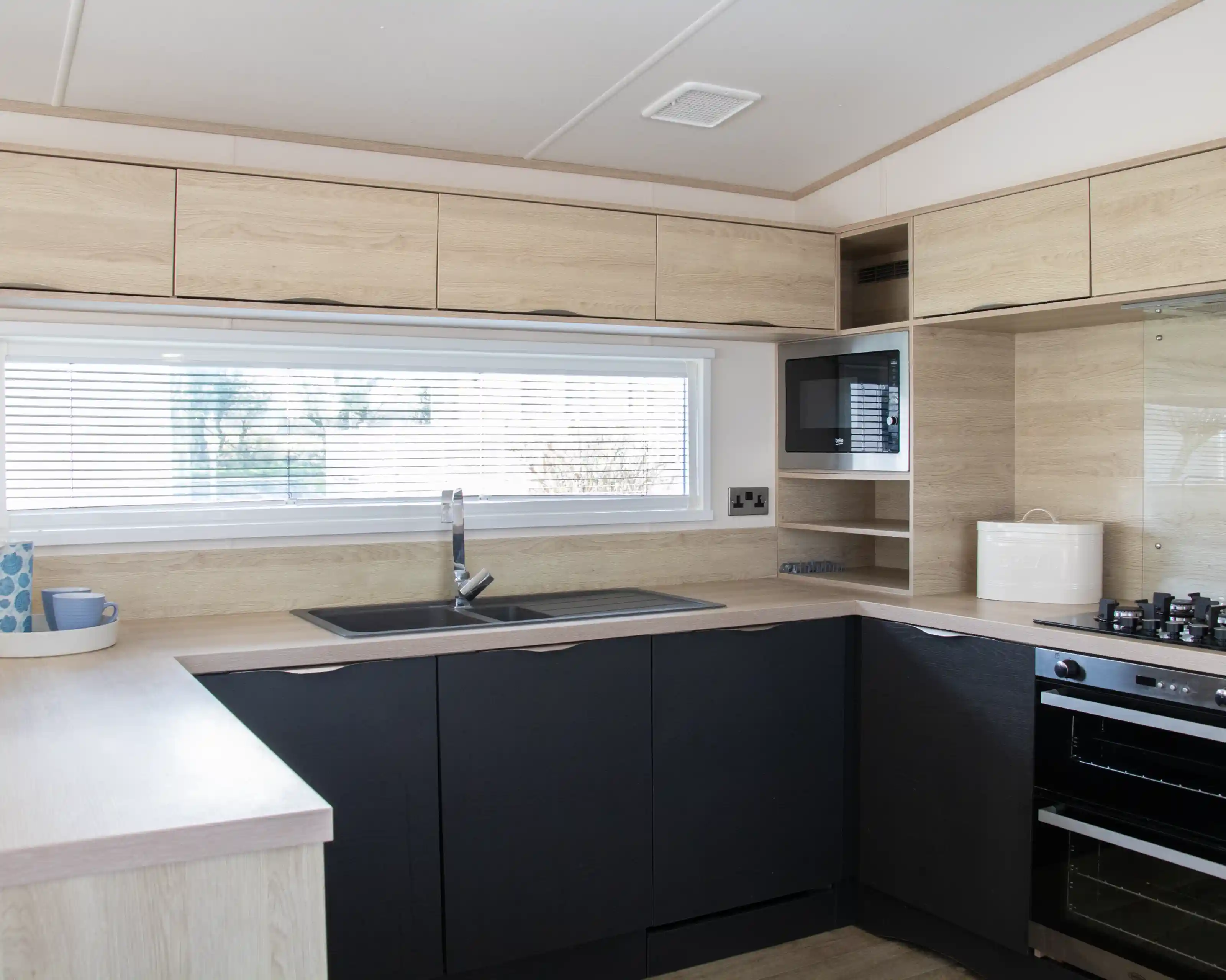 Modern kitchen featuring wooden cabinetry in light and dark tones. A large window with blinds provides natural light. The layout includes a sink, stovetop, and microwave, with minimalistic shelving above.