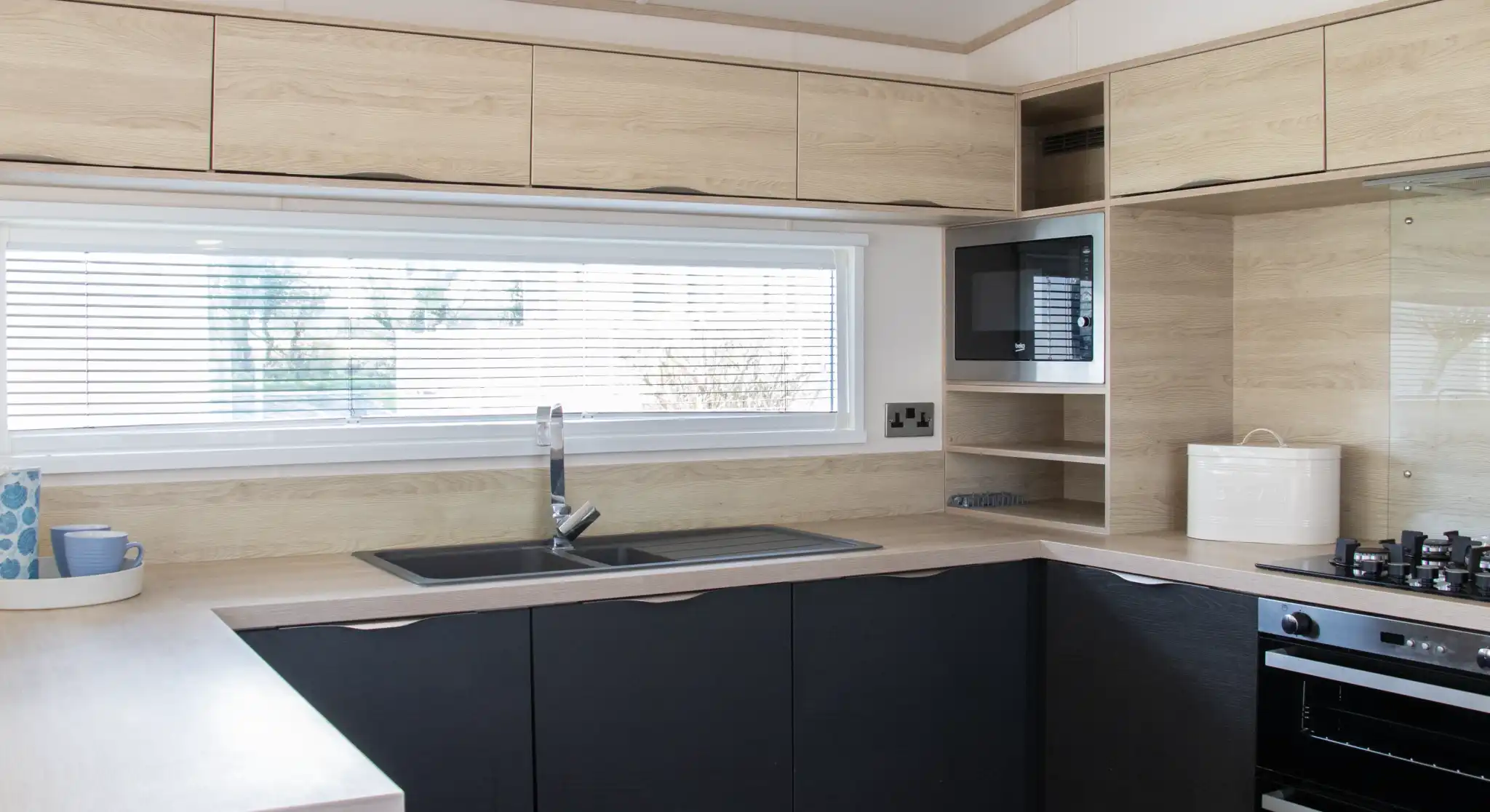 Modern kitchen featuring wooden cabinetry in light and dark tones. A large window with blinds provides natural light. The layout includes a sink, stovetop, and microwave, with minimalistic shelving above.
