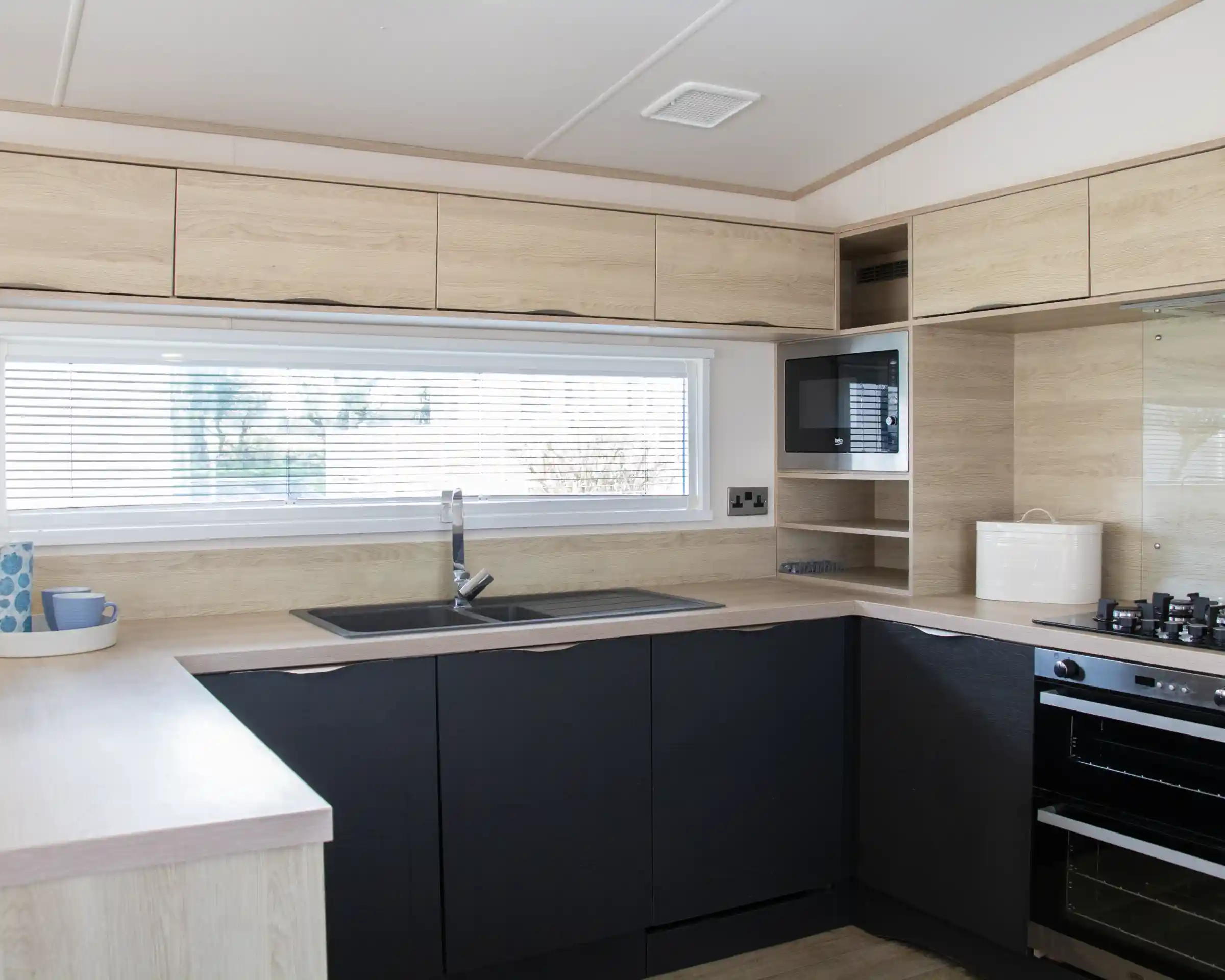 Modern kitchen featuring wooden cabinetry in light and dark tones. A large window with blinds provides natural light. The layout includes a sink, stovetop, and microwave, with minimalistic shelving above.