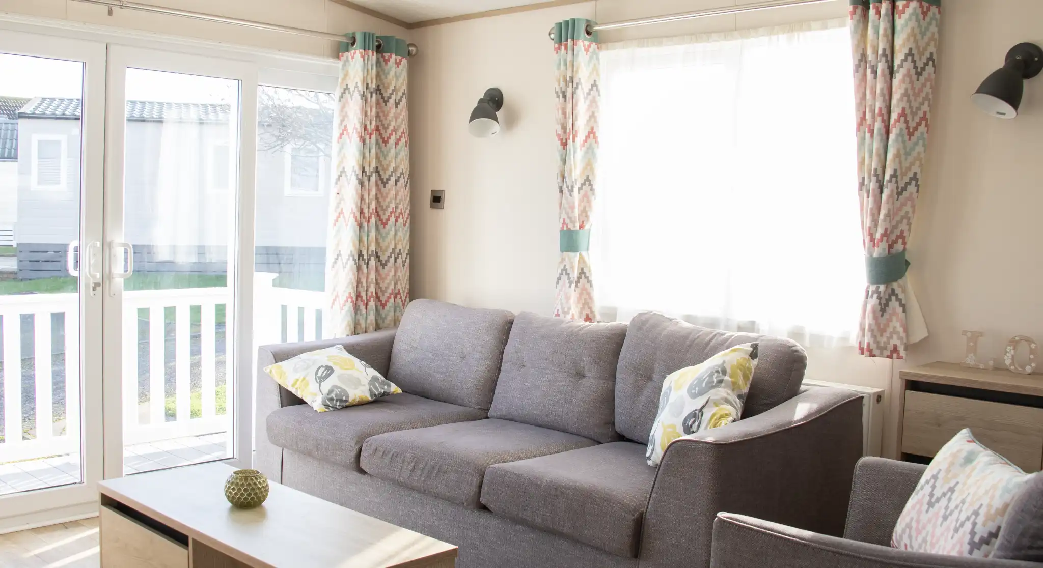 A bright and cozy living room features a gray sofa with patterned cushions, a wooden coffee table, and colorful curtains. Large glass doors provide natural light and lead to an outdoor area.