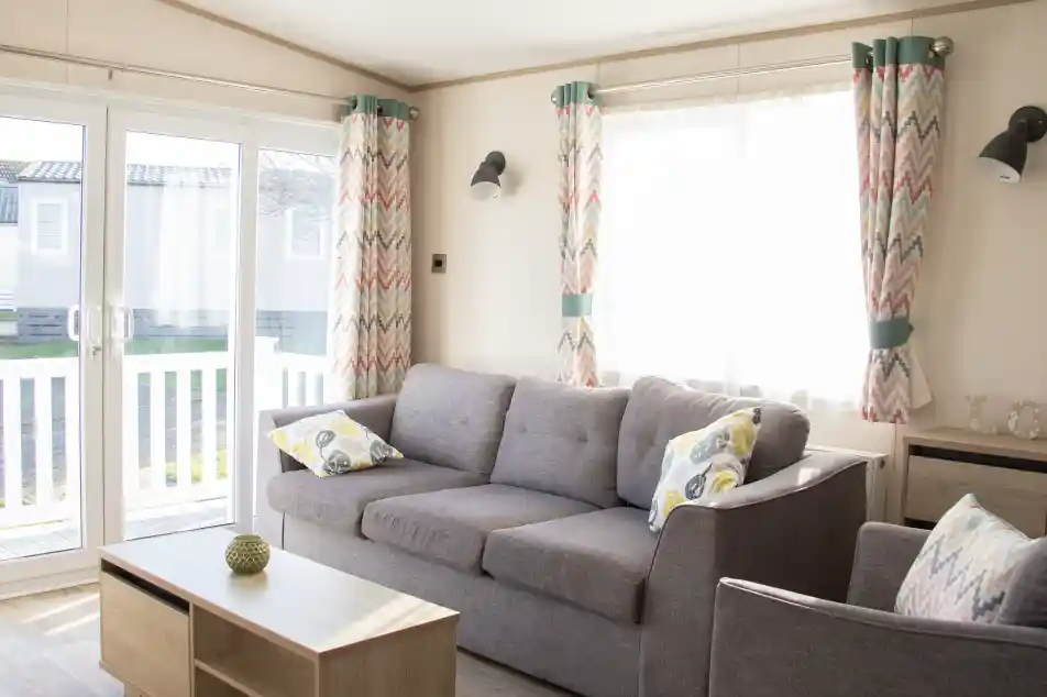 A bright and cozy living room features a gray sofa with patterned cushions, a wooden coffee table, and colorful curtains. Large glass doors provide natural light and lead to an outdoor area.