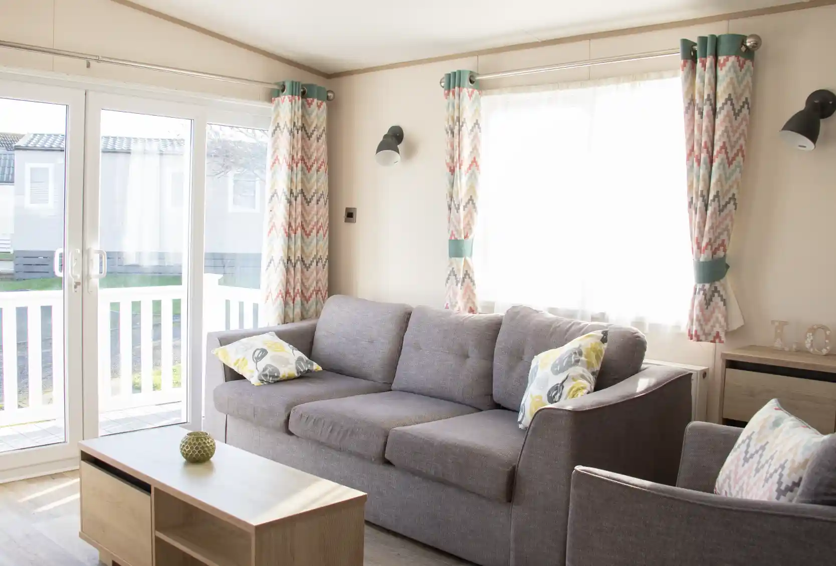A bright and cozy living room features a gray sofa with patterned cushions, a wooden coffee table, and colorful curtains. Large glass doors provide natural light and lead to an outdoor area.