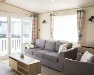 A bright and cozy living room features a gray sofa with patterned cushions, a wooden coffee table, and colorful curtains. Large glass doors provide natural light and lead to an outdoor area.