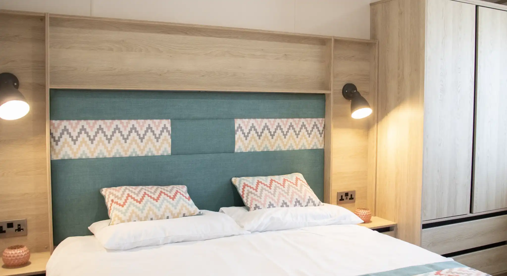 A cozy bedroom featuring a neatly made bed with patterned pillows and a colorful bedspread. The headboard is upholstered in teal with a geometric design, and there are wooden cabinets and bedside lamps in a warm, modern style.