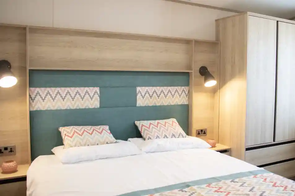 A cozy bedroom featuring a neatly made bed with patterned pillows and a colorful bedspread. The headboard is upholstered in teal with a geometric design, and there are wooden cabinets and bedside lamps in a warm, modern style.