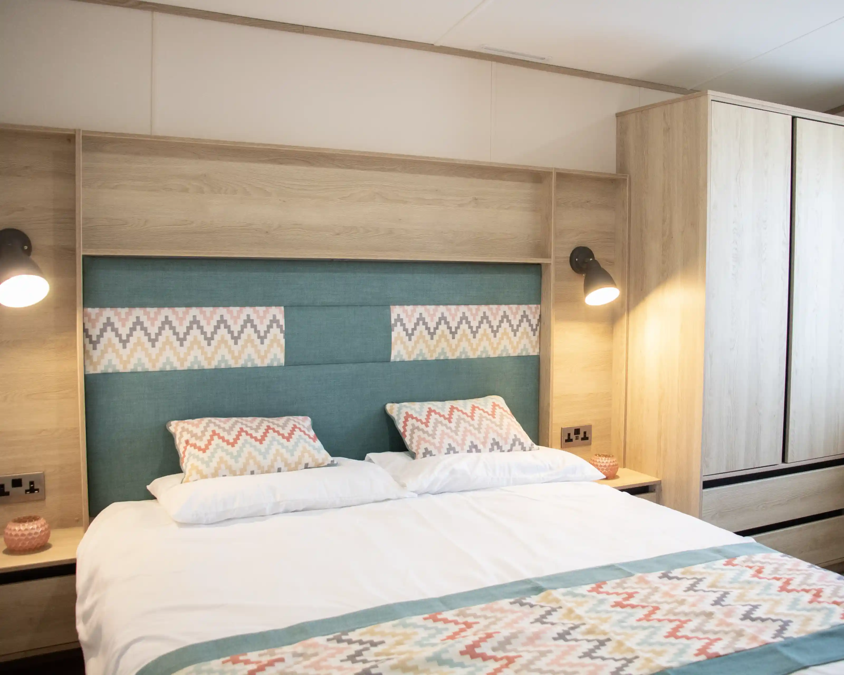 A cozy bedroom featuring a neatly made bed with patterned pillows and a colorful bedspread. The headboard is upholstered in teal with a geometric design, and there are wooden cabinets and bedside lamps in a warm, modern style.