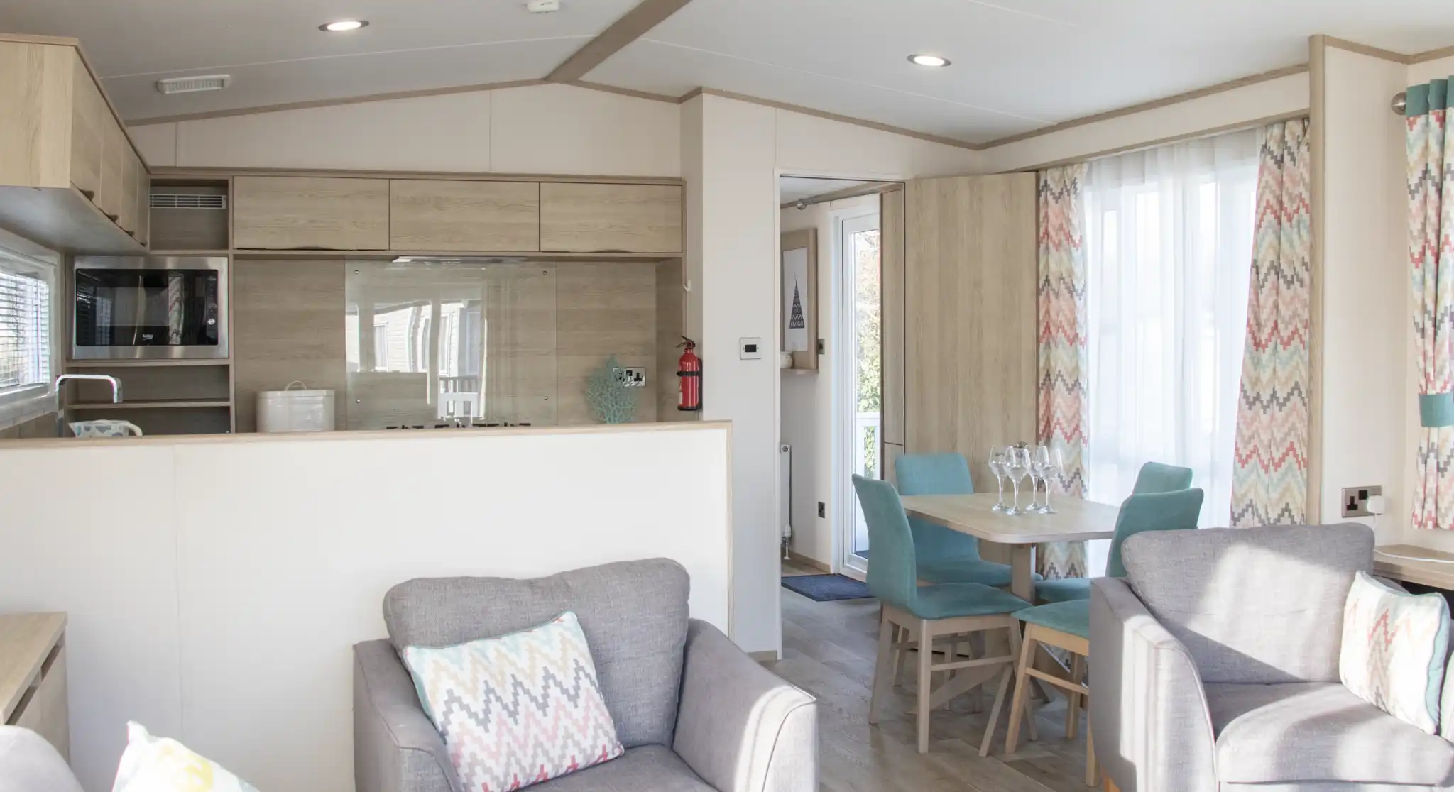 A modern, cozy interior of a holiday home featuring a kitchenette with wood accents, a dining area with a turquoise table and chairs, and comfortable gray armchairs with decorative pillows. Natural light streams through large windows with colorful, patterned curtains.