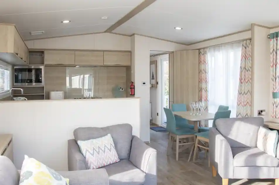 A modern, cozy interior of a holiday home featuring a kitchenette with wood accents, a dining area with a turquoise table and chairs, and comfortable gray armchairs with decorative pillows. Natural light streams through large windows with colorful, patterned curtains.