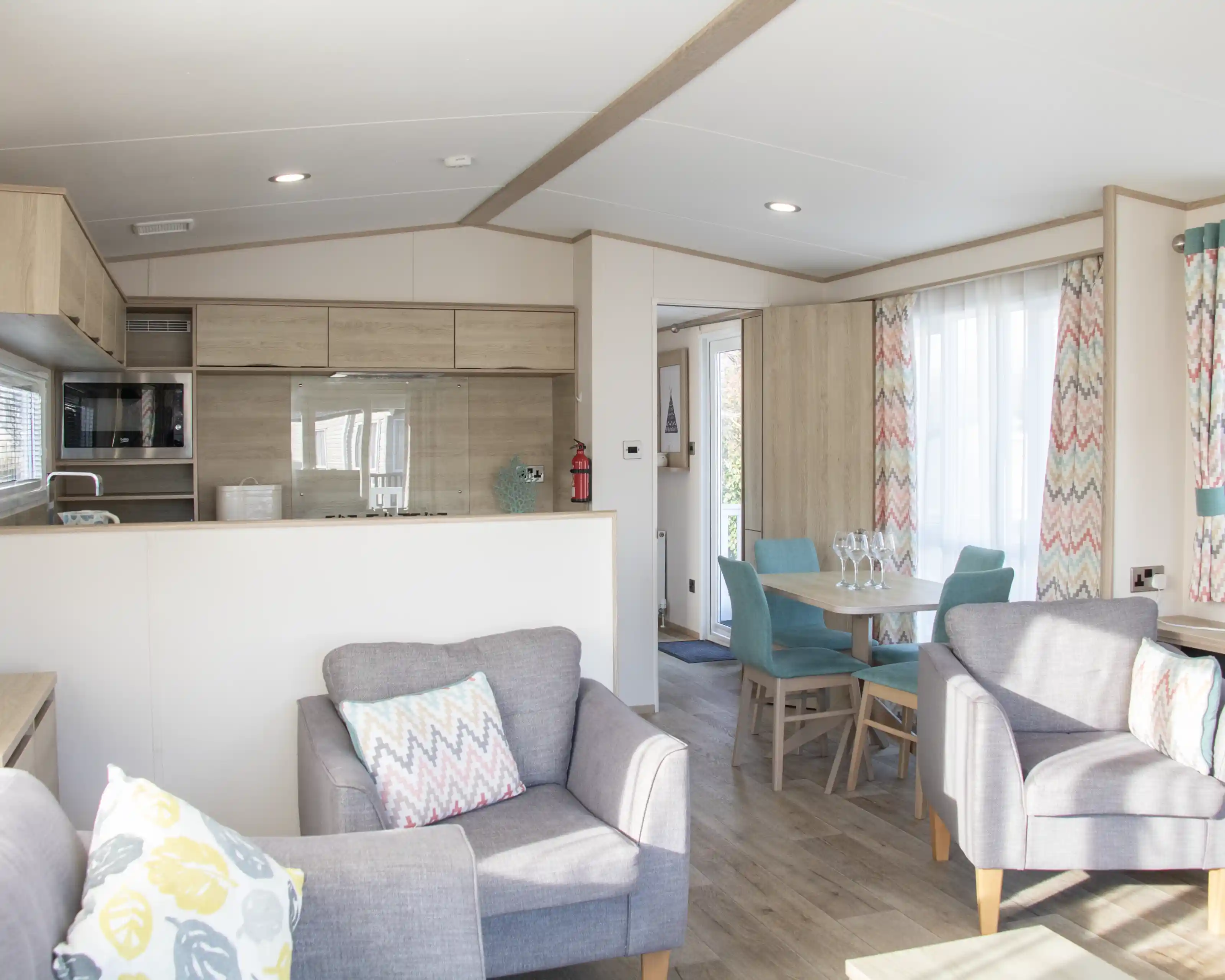 A modern, cozy interior of a holiday home featuring a kitchenette with wood accents, a dining area with a turquoise table and chairs, and comfortable gray armchairs with decorative pillows. Natural light streams through large windows with colorful, patterned curtains.