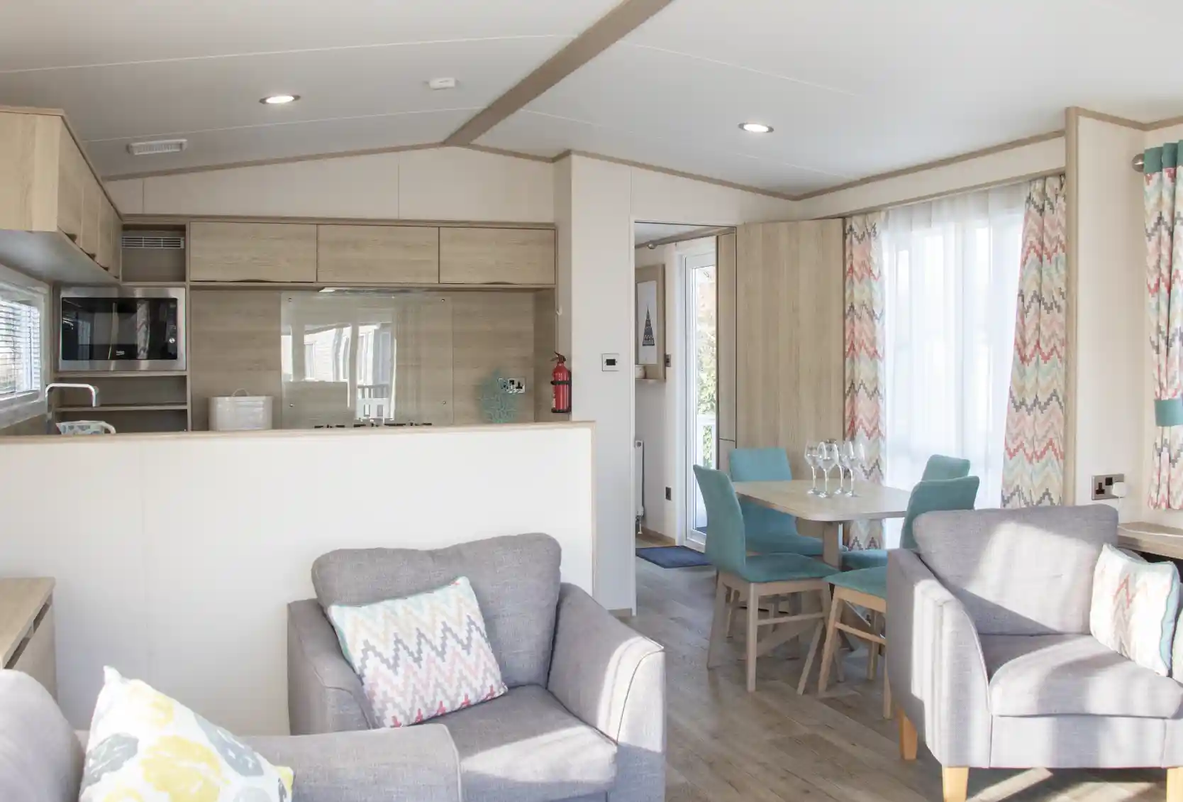 A modern, cozy interior of a holiday home featuring a kitchenette with wood accents, a dining area with a turquoise table and chairs, and comfortable gray armchairs with decorative pillows. Natural light streams through large windows with colorful, patterned curtains.