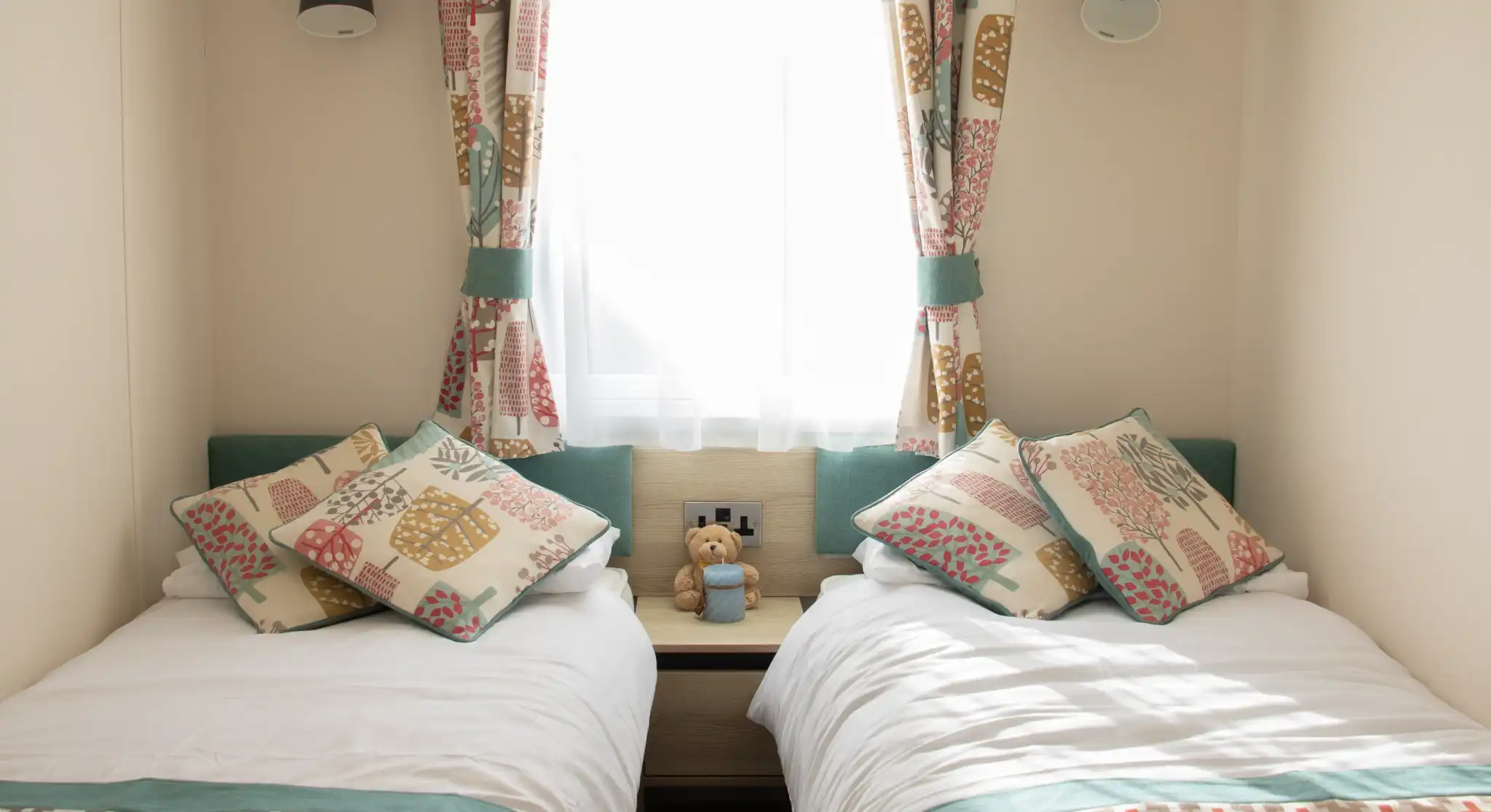 A cozy bedroom featuring two single beds with colorful patterned pillows. A teddy bear sits on a nightstand between the beds. Natural light streams in through a window dressed with light curtains.
