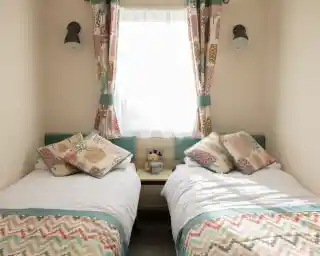 A cozy bedroom featuring two single beds with colorful patterned pillows. A teddy bear sits on a nightstand between the beds. Natural light streams in through a window dressed with light curtains.