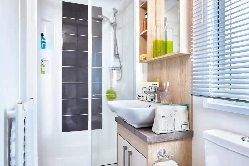 A modern bathroom featuring a glass shower enclosure, a sleek sink with decorative bottles and toiletries on shelves, a countertop, and a toilet. Natural light filters through the blinds.