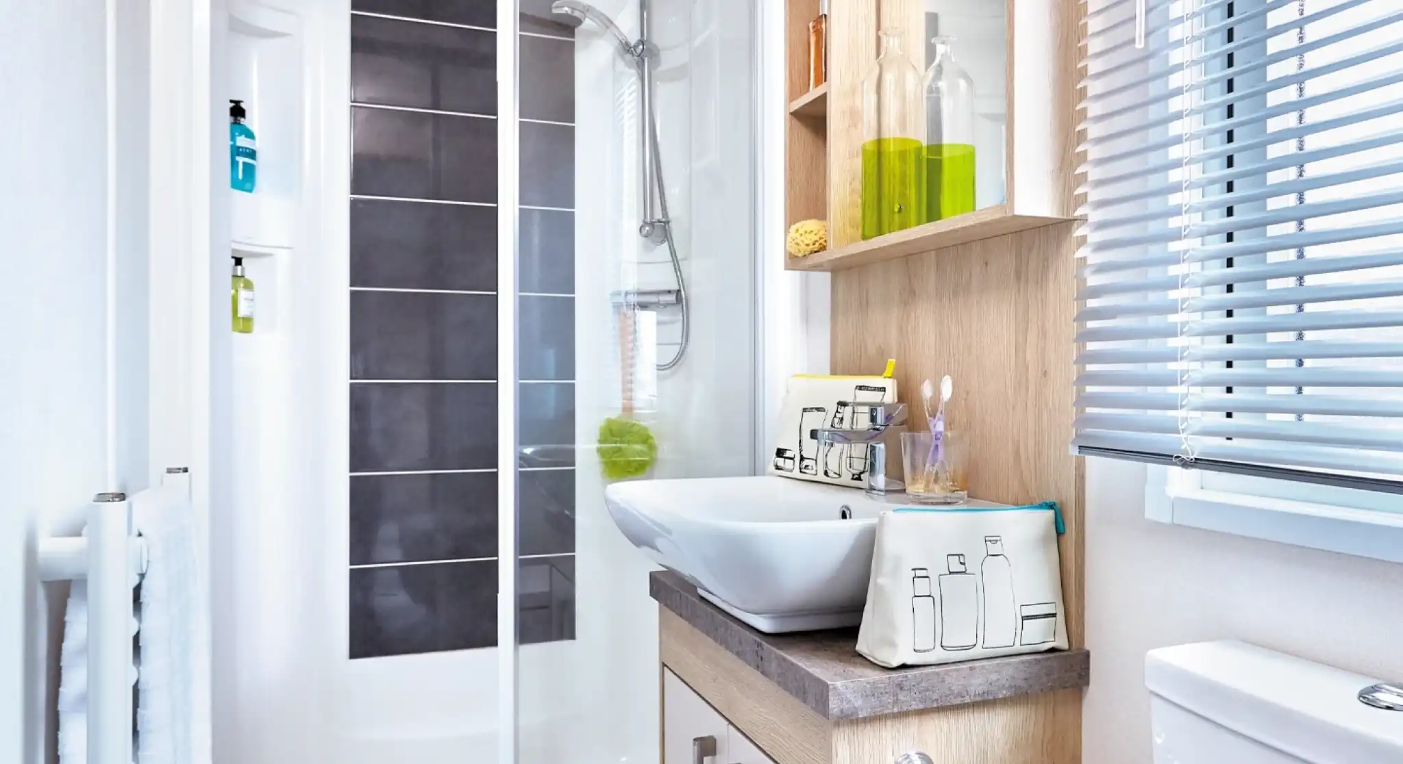 A modern bathroom featuring a glass shower enclosure, a sleek sink with decorative bottles and toiletries on shelves, a countertop, and a toilet. Natural light filters through the blinds.