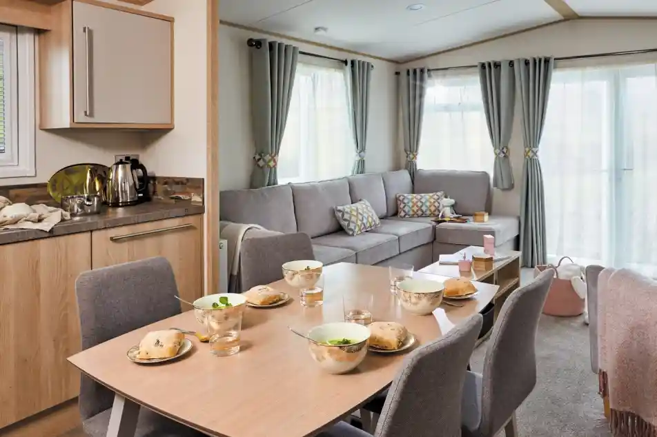 A cozy living space featuring a dining table set for breakfast with bowls and glasses. A comfortable gray sofa is positioned nearby, complemented by colorful cushions. Large windows allow natural light to brighten the room, which has a warm and inviting atmosphere.