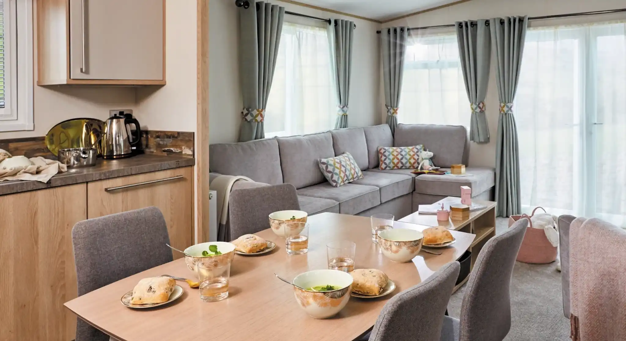 A cozy living space featuring a dining table set for breakfast with bowls and glasses. A comfortable gray sofa is positioned nearby, complemented by colorful cushions. Large windows allow natural light to brighten the room, which has a warm and inviting atmosphere.