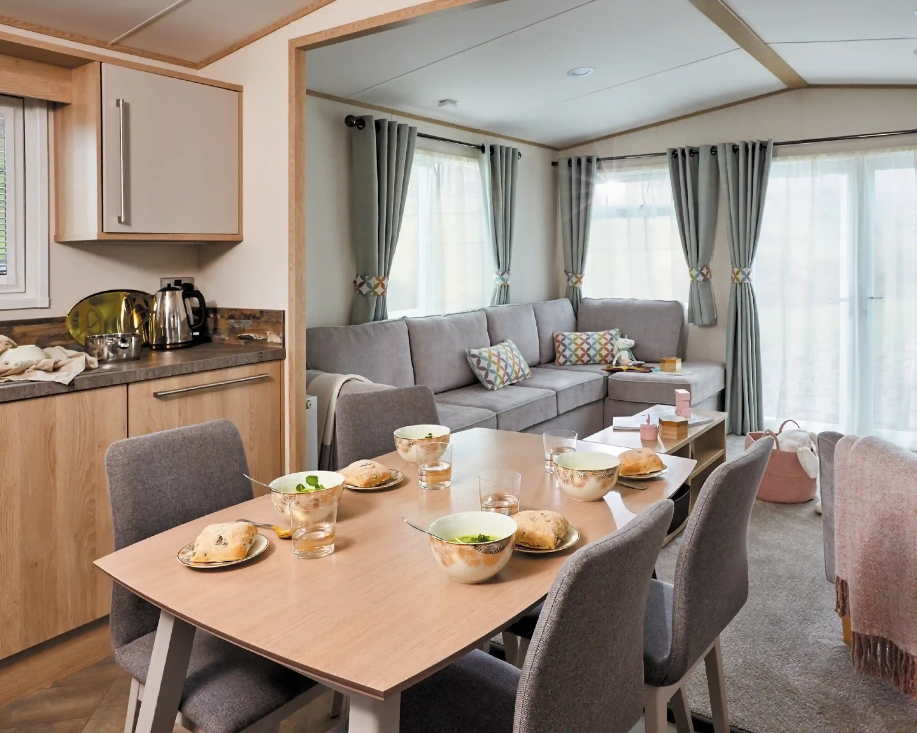 A cozy living space featuring a dining table set for breakfast with bowls and glasses. A comfortable gray sofa is positioned nearby, complemented by colorful cushions. Large windows allow natural light to brighten the room, which has a warm and inviting atmosphere.