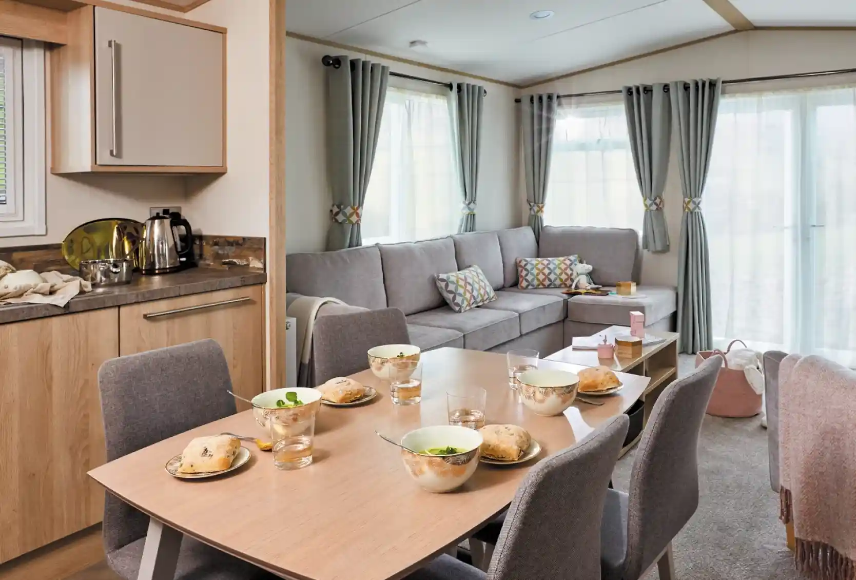 A cozy living space featuring a dining table set for breakfast with bowls and glasses. A comfortable gray sofa is positioned nearby, complemented by colorful cushions. Large windows allow natural light to brighten the room, which has a warm and inviting atmosphere.