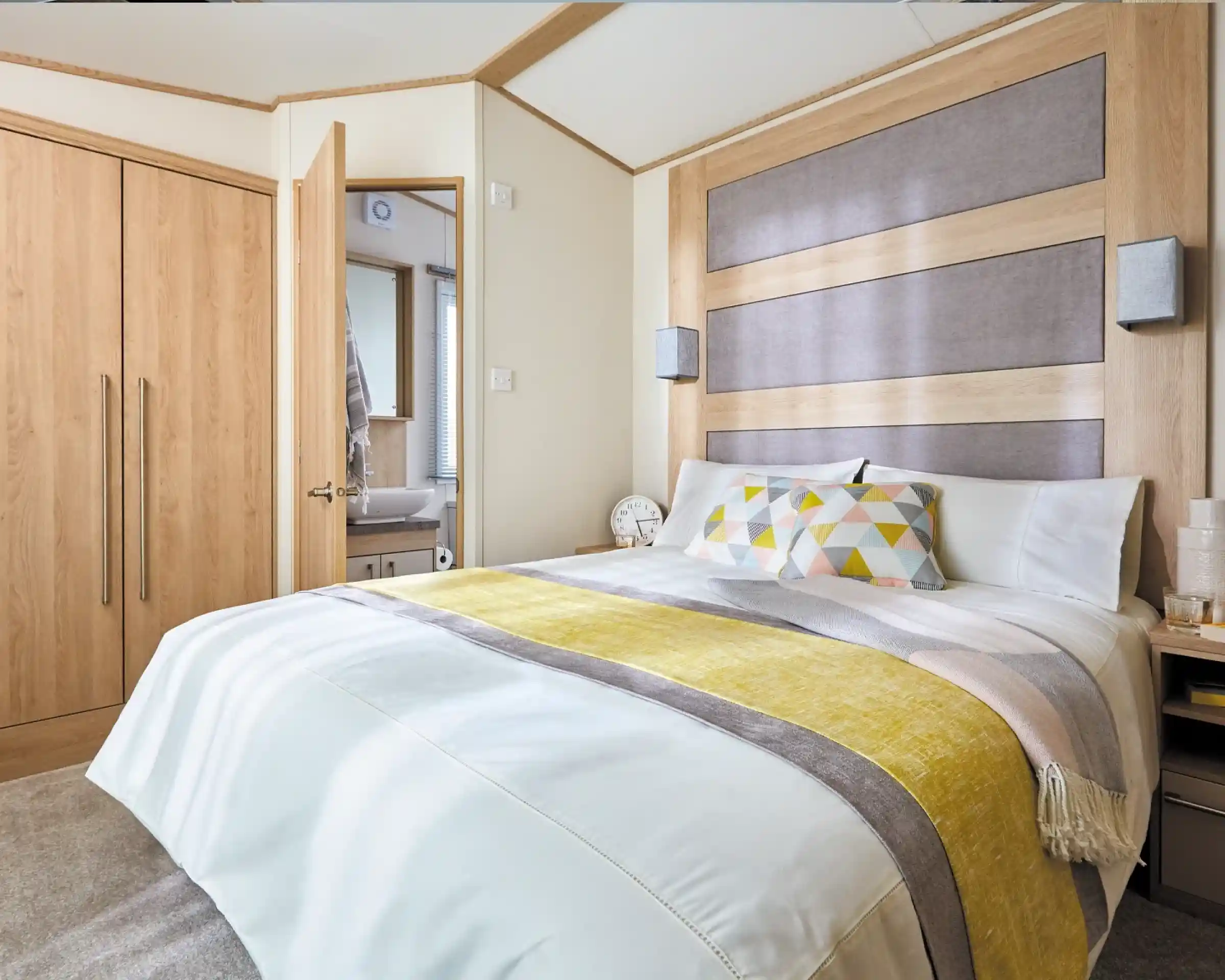A modern bedroom with a large bed featuring a white comforter and colorful patterned pillows. The headboard is made of wood with a gray panel, and there are stylish bedside lamps. A closet is visible on the left, and a bathroom can be seen through an open door in the background.