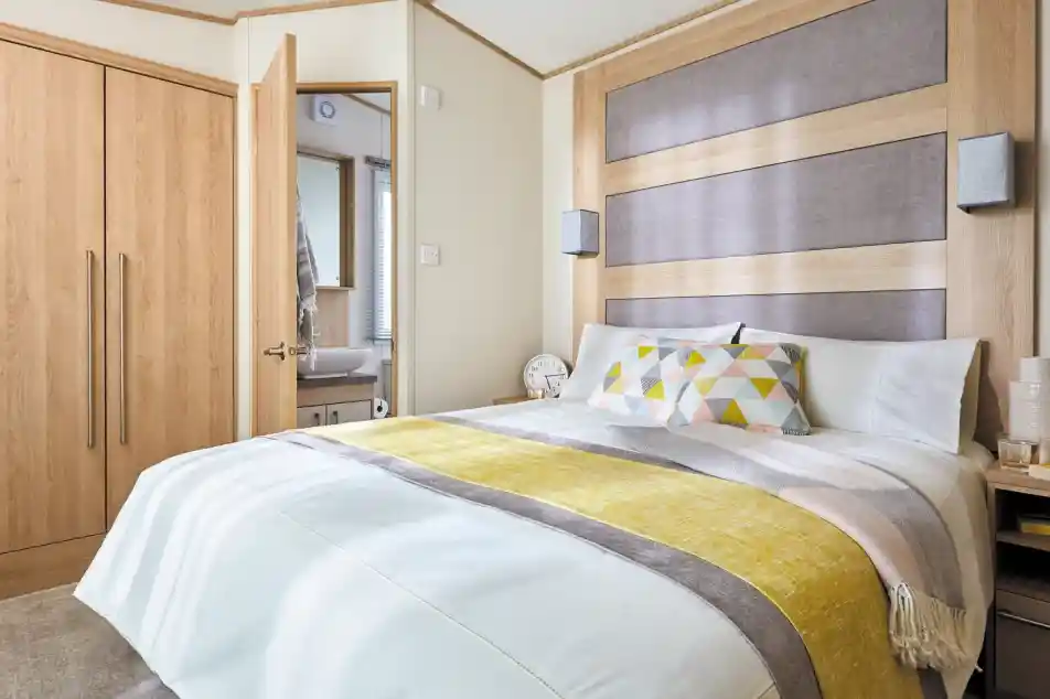A modern bedroom with a large bed featuring a white comforter and colorful patterned pillows. The headboard is made of wood with a gray panel, and there are stylish bedside lamps. A closet is visible on the left, and a bathroom can be seen through an open door in the background.