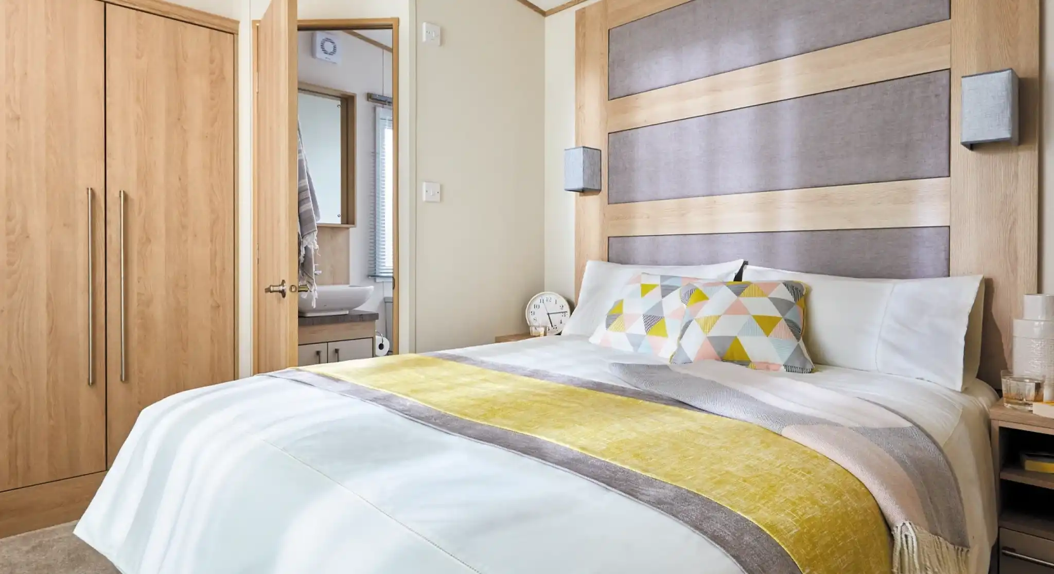 A modern bedroom with a large bed featuring a white comforter and colorful patterned pillows. The headboard is made of wood with a gray panel, and there are stylish bedside lamps. A closet is visible on the left, and a bathroom can be seen through an open door in the background.