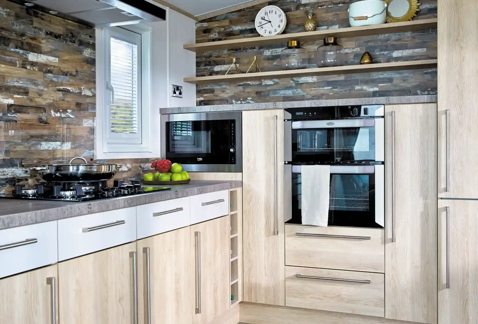 A modern kitchen features light wood cabinetry with a gray countertop. On the left, there's a gas stove with a frying pan, while above it hangs a sleek black microwave. Two built-in ovens are topped with a white towel and flanked by decorative items on open shelves. The wall has a stylish stone pattern, and a round clock is mounted above the appliances. Green apples are displayed on the counter.