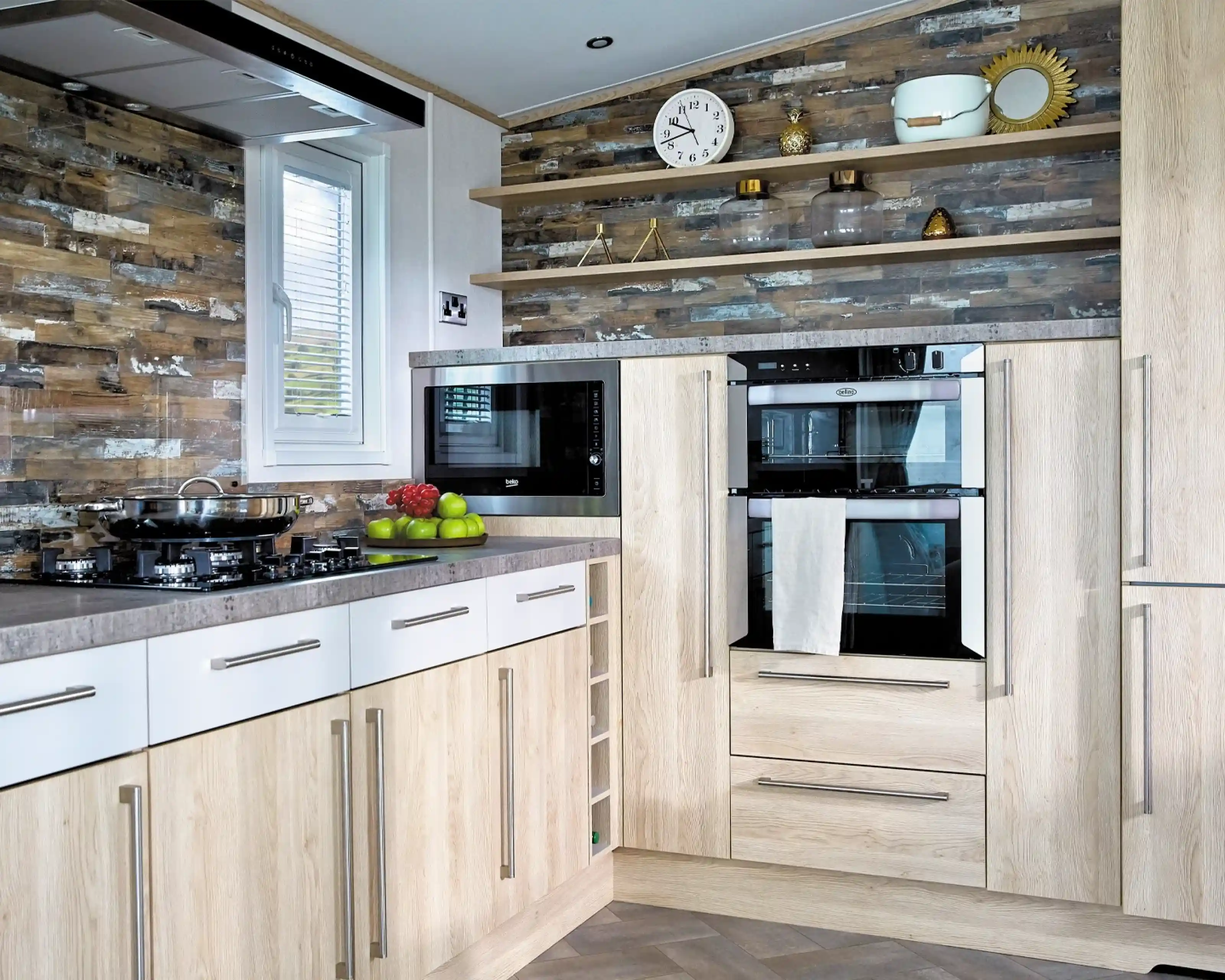 A modern kitchen features light wood cabinetry with a gray countertop. On the left, there's a gas stove with a frying pan, while above it hangs a sleek black microwave. Two built-in ovens are topped with a white towel and flanked by decorative items on open shelves. The wall has a stylish stone pattern, and a round clock is mounted above the appliances. Green apples are displayed on the counter.