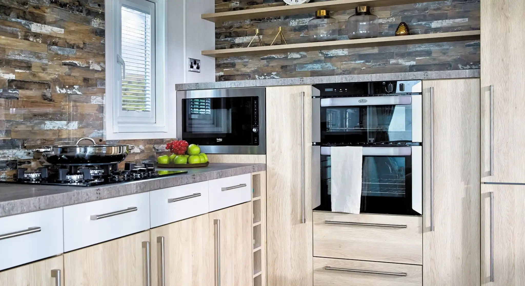 A modern kitchen features light wood cabinetry with a gray countertop. On the left, there's a gas stove with a frying pan, while above it hangs a sleek black microwave. Two built-in ovens are topped with a white towel and flanked by decorative items on open shelves. The wall has a stylish stone pattern, and a round clock is mounted above the appliances. Green apples are displayed on the counter.