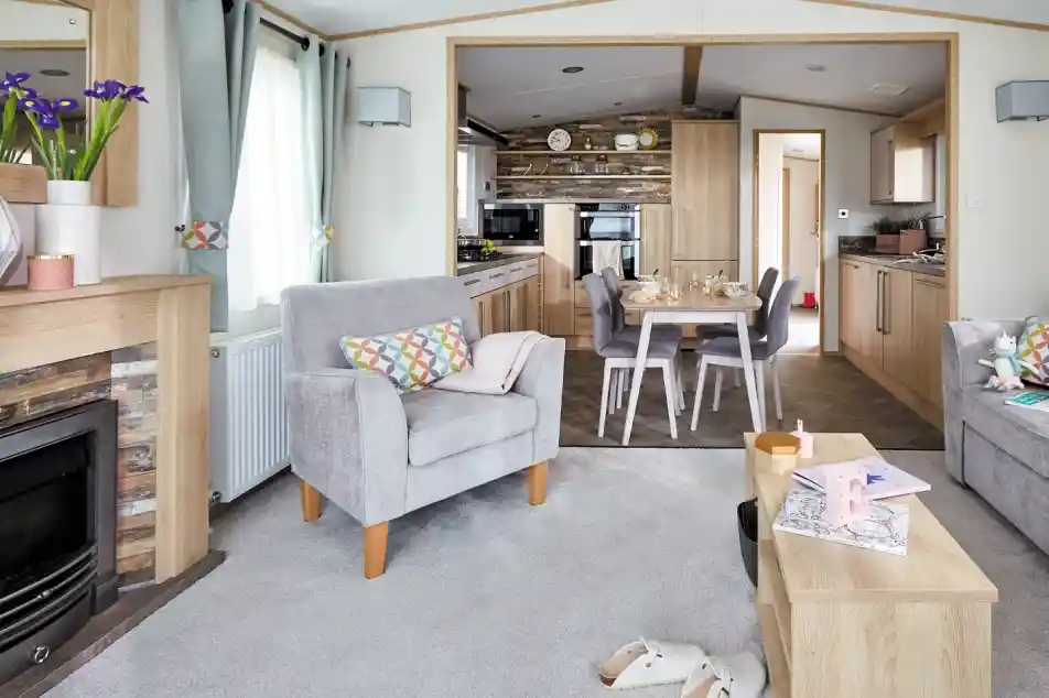 A cozy living room features two gray armchairs with colorful cushions, a wooden coffee table, and a modern electric fireplace. In the background, a well-equipped kitchen is visible, with wooden cabinetry and a dining area set for a meal. Light filters through large windows, adding a warm ambiance to the space.